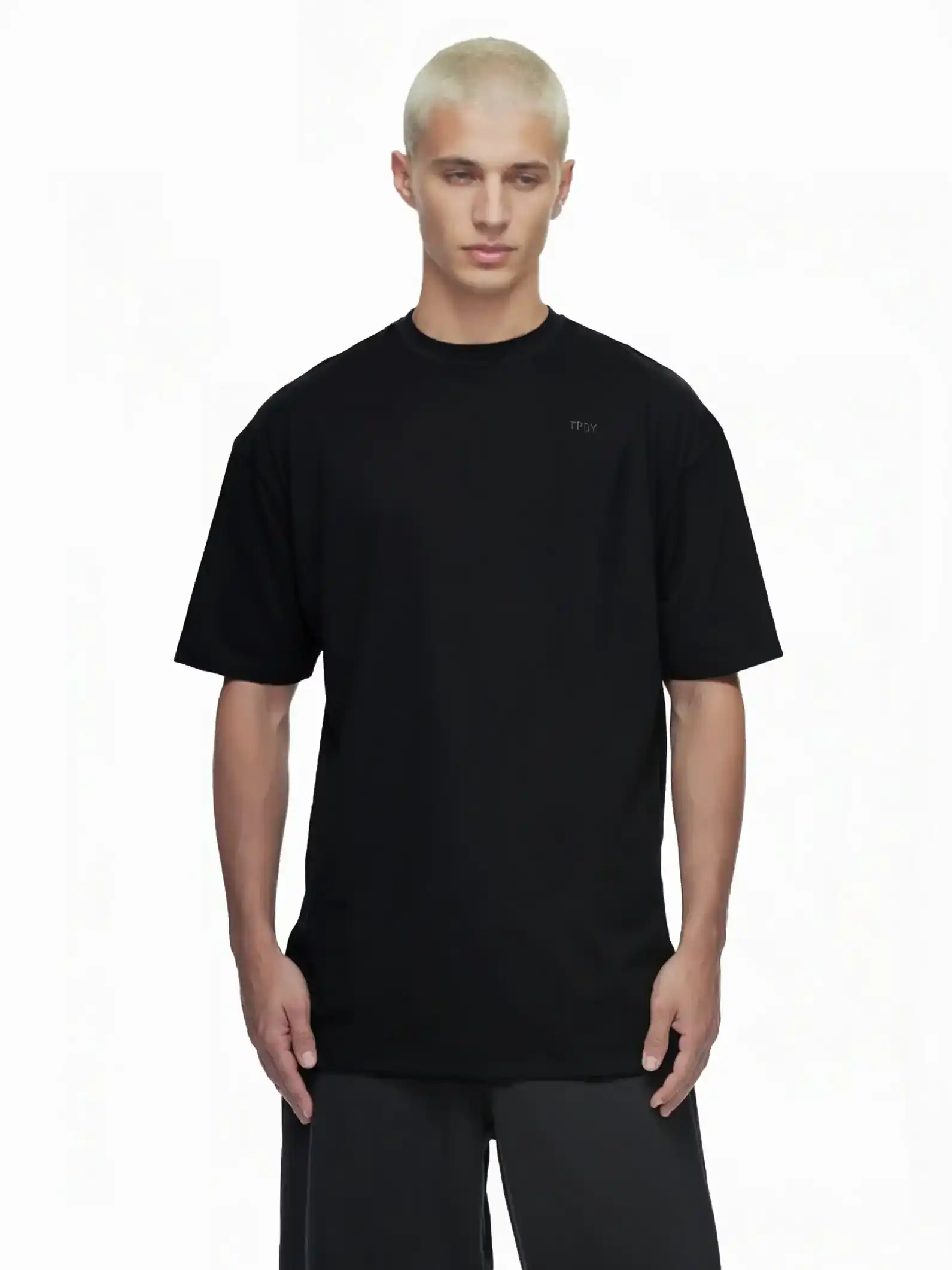 Eine Person mit kurzen, gebleichten blonden Haaren trägt das trueprodigy Haram Oversize-Herren-T-Shirt mit dezentem TPDY-Sticklogo in Schwarz und schwarzer Hose vor weißem Hintergrund. Das Shirt hat eine dezente Bruststickerei.