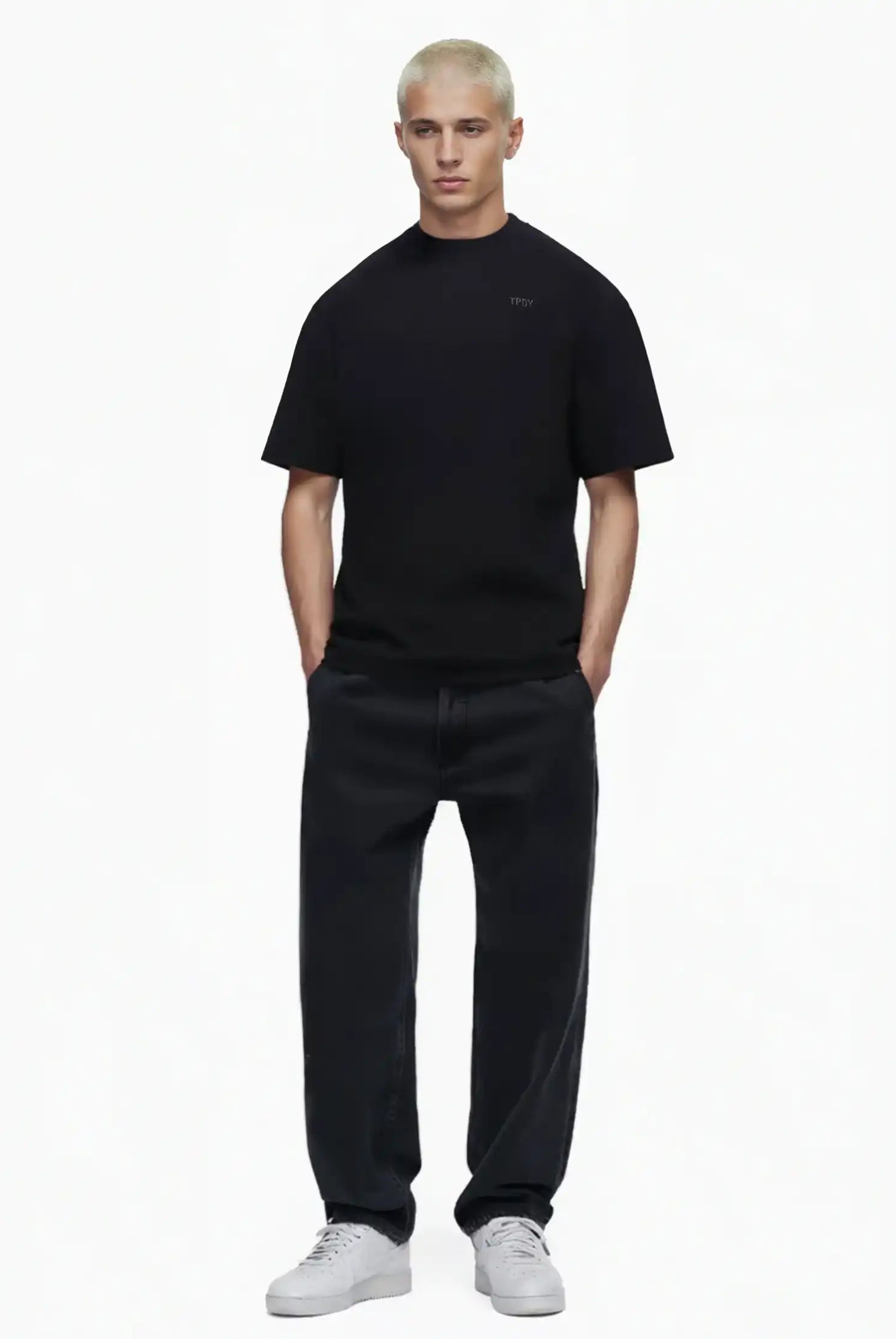 Eine Person mit kurzen, gebleichten blonden Haaren trägt das Trueprodigy Haram Oversize-Herren-T-Shirt mit dezentem TPDY-Sticklogo in Schwarz, weite schwarze Jeans und weiße Turnschuhe. Sie steht mit den Händen in den Taschen vor einem schlichten weißen Hintergrund.