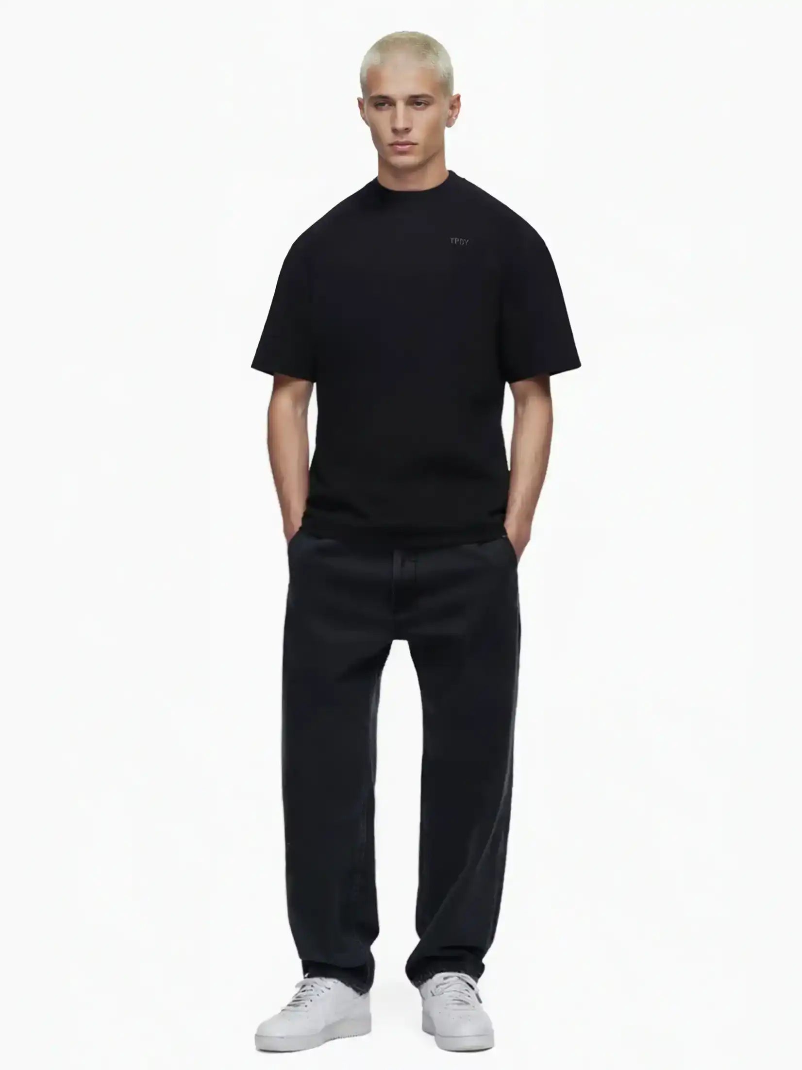 Eine Person mit kurzen, gebleichten blonden Haaren trägt das Trueprodigy Haram Oversize-Herren-T-Shirt mit dezentem TPDY-Sticklogo in Schwarz, weite schwarze Jeans und weiße Turnschuhe. Sie steht mit den Händen in den Taschen vor einem schlichten weißen Hintergrund.