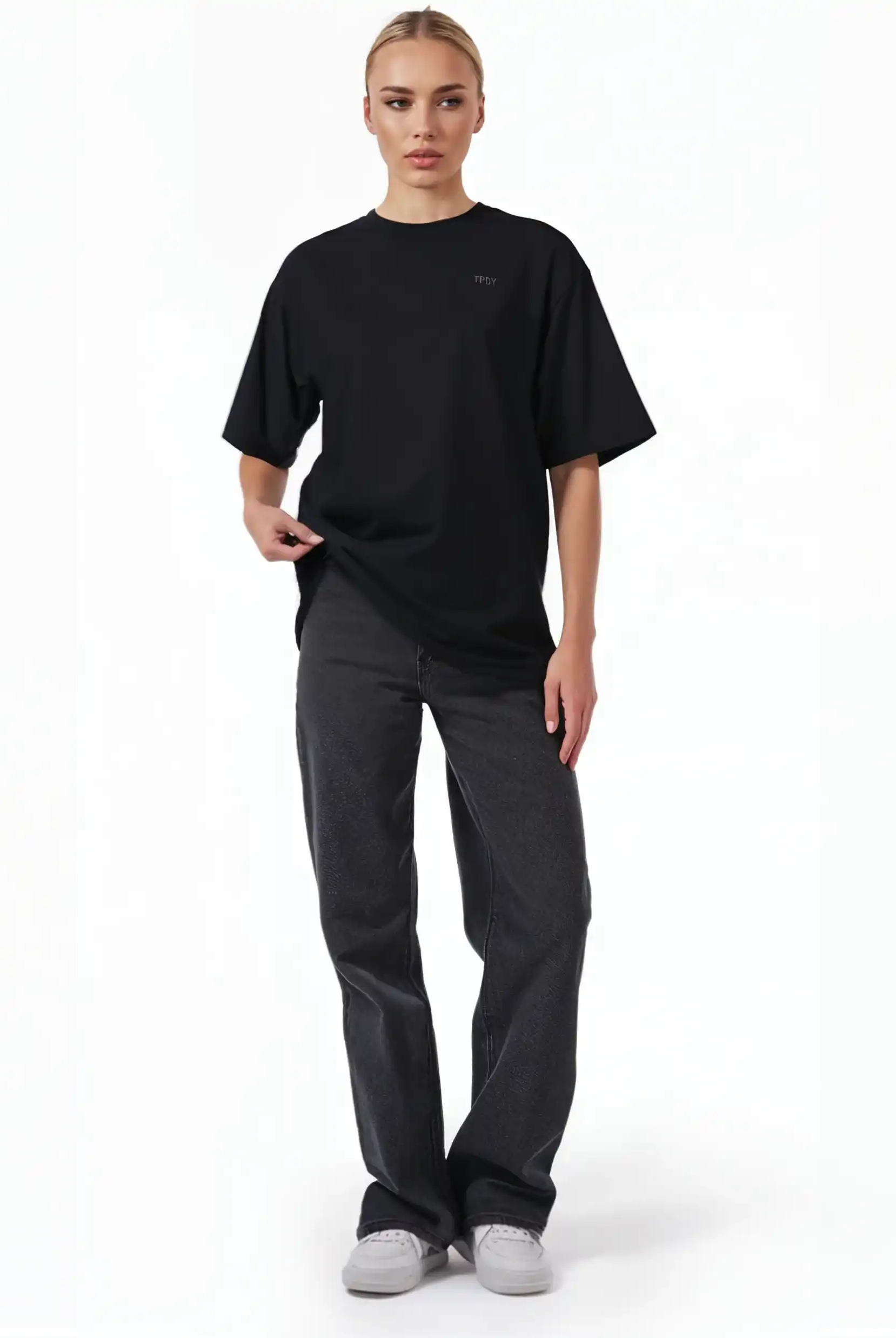 Eine Person steht mit dem Gesicht nach vorne vor einem weißen Hintergrund und trägt das trueprodigy Haram F Oversize-T-Shirt mit TPDY-Stickerei & cleaner Optik, dunkle Jeans mit geradem Bein und weiße Turnschuhe. Eine Hand zieht leicht an der Seite des Unisex-T-Shirts.