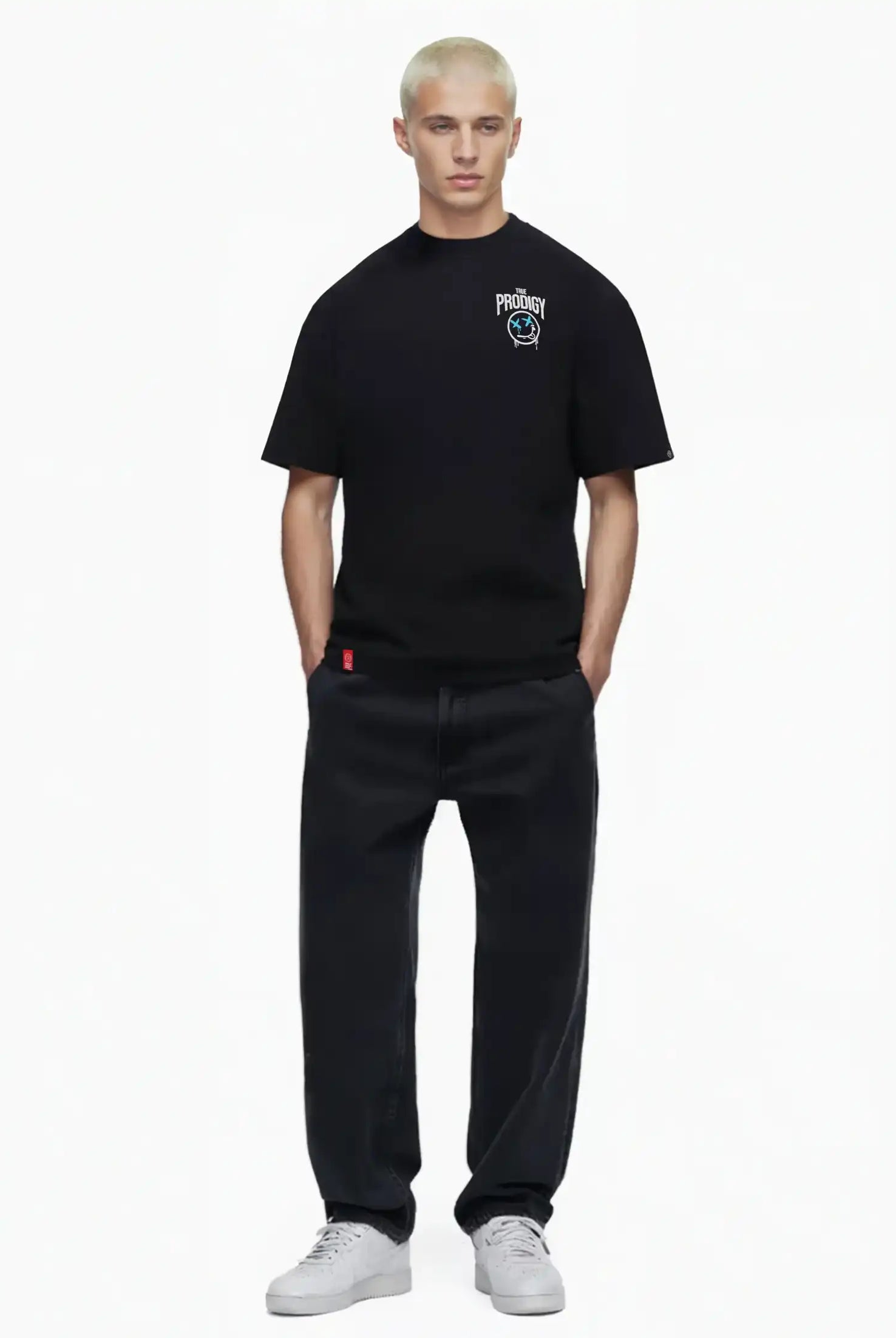 Eine Person mit kurzen, gebleichten blonden Haaren modelliert das trueprodigy Kenji Oversize-T-Shirt mit Brustprint und großem Rückenmotiv, gepaart mit lockeren schwarzen Jeans und weißen Turnschuhen, vor einem schlichten weißen Hintergrund.