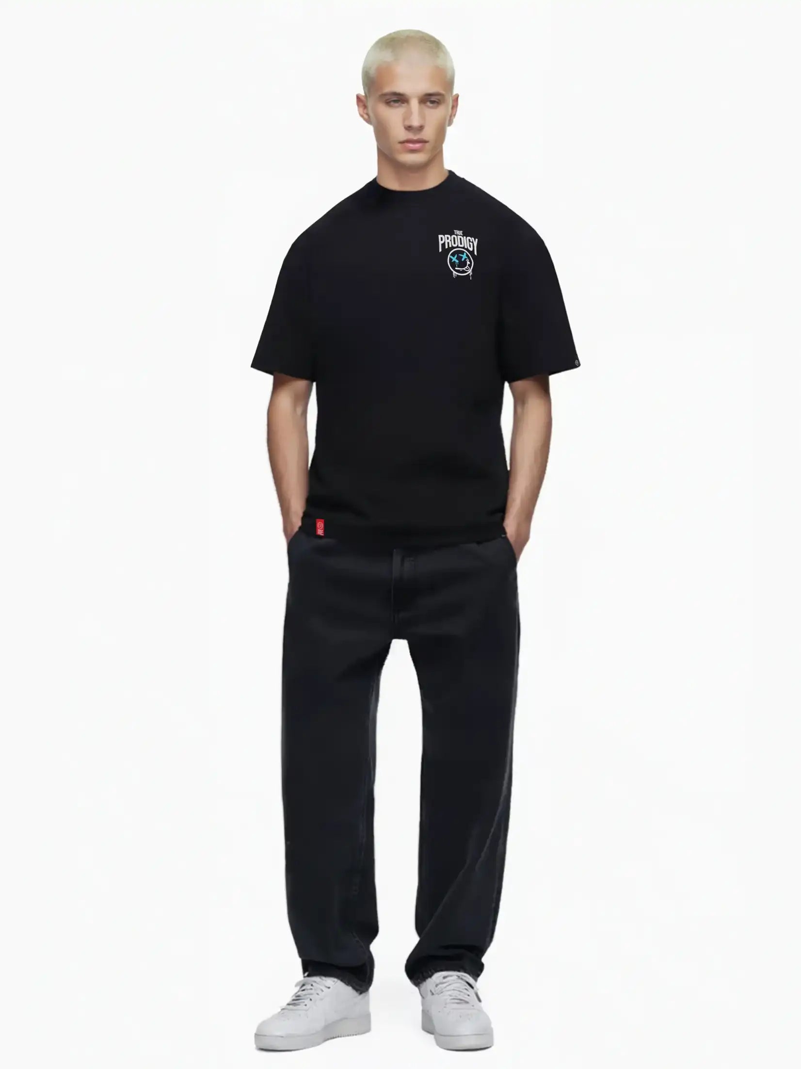 Eine Person mit kurzen, gebleichten blonden Haaren modelliert das trueprodigy Kenji Oversize-T-Shirt mit Brustprint und großem Rückenmotiv, gepaart mit lockeren schwarzen Jeans und weißen Turnschuhen, vor einem schlichten weißen Hintergrund.