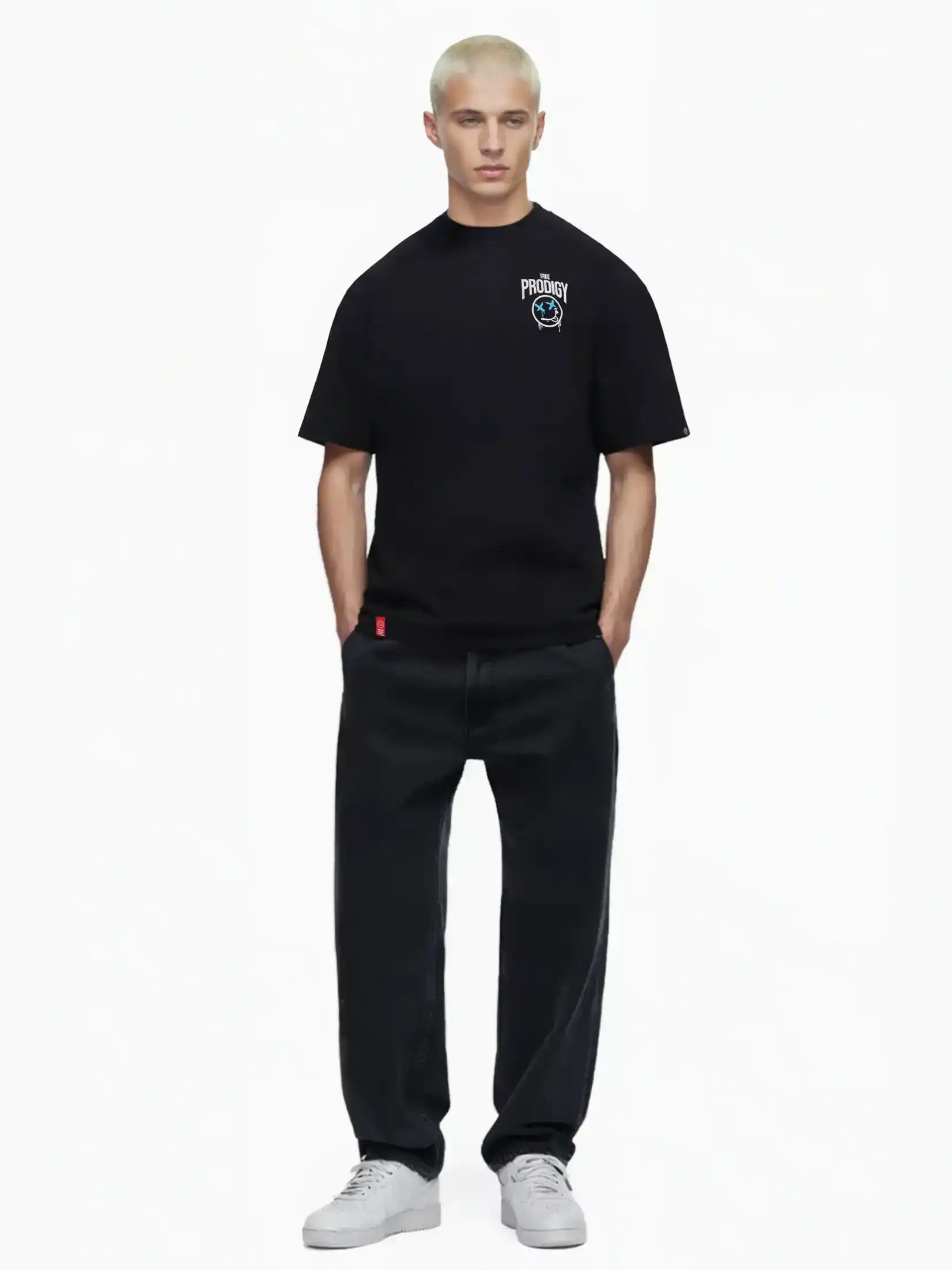 Eine Person mit kurzen, gebleichten blonden Haaren modelliert das trueprodigy Kenji Oversize-T-Shirt mit Brustprint und großem Rückenmotiv, gepaart mit lockeren schwarzen Jeans und weißen Turnschuhen, vor einem schlichten weißen Hintergrund.