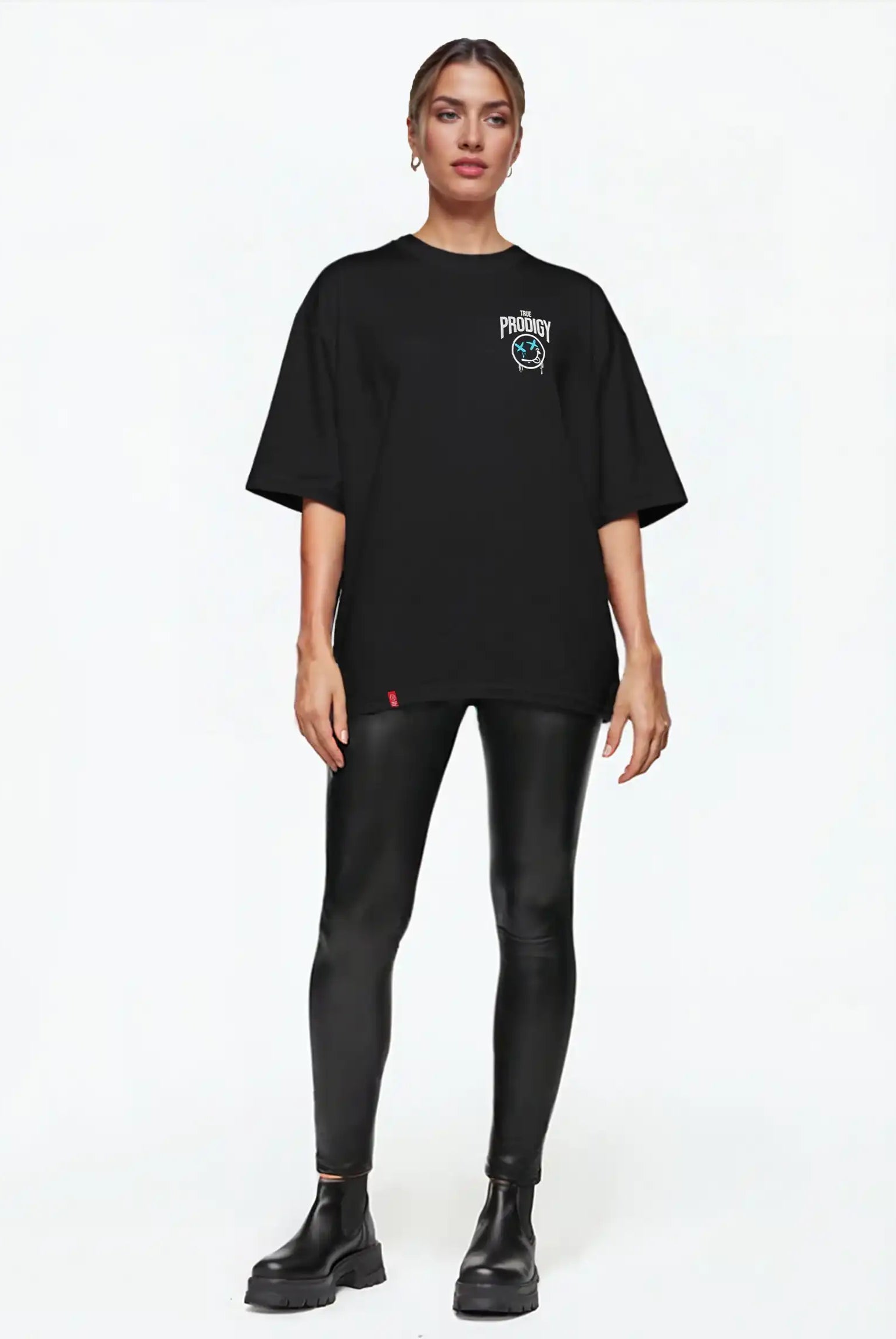 Eine Person steht vor einem schlichten weißen Hintergrund und trägt das trueprodigy Kenji F Oversize-Damen-T-Shirt mit Graffiti-Brust- und Rückenprint, gepaart mit glänzenden schwarzen Leggings und schwarzen Ankle Boots.