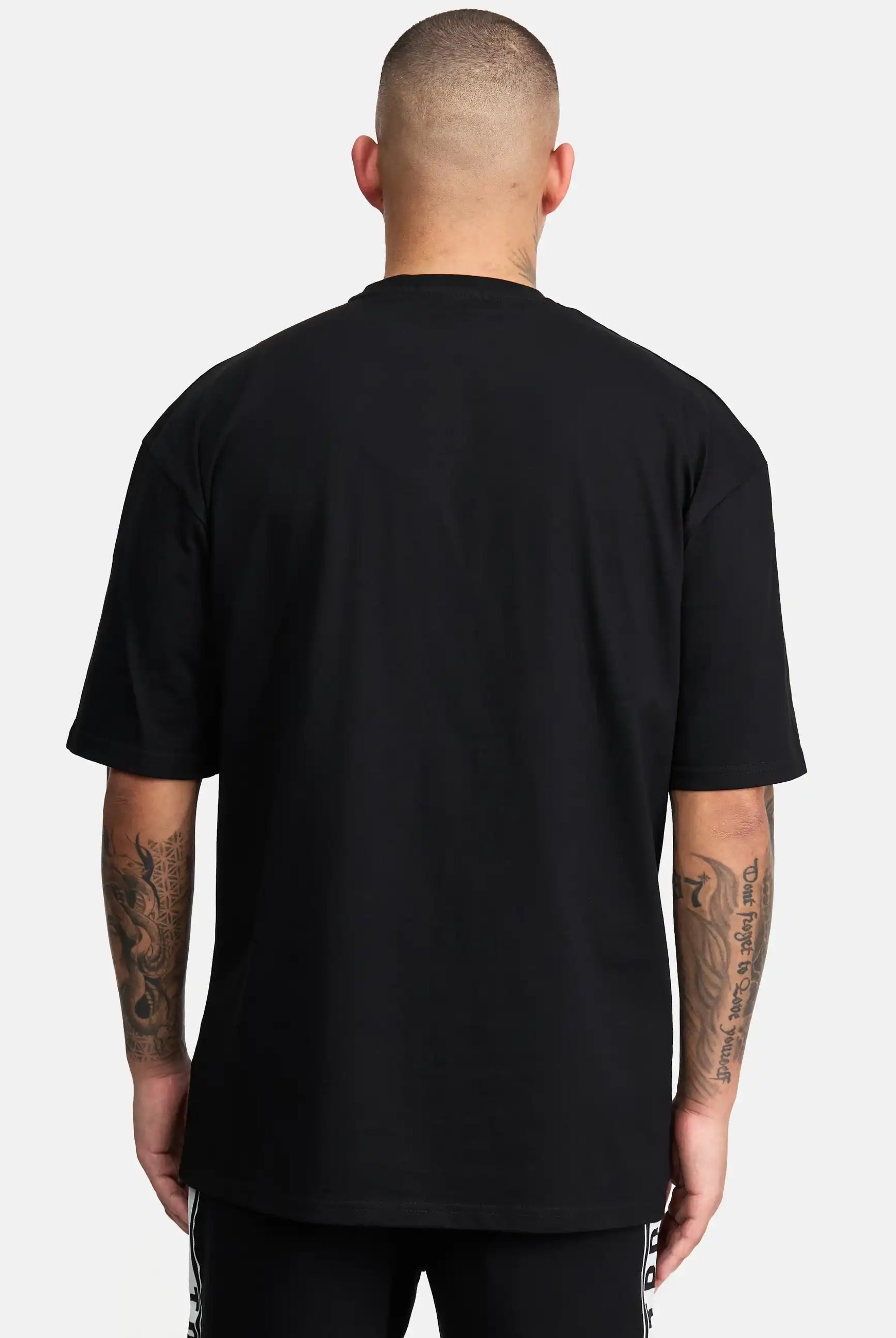 Gezeigt wird eine tätowierte Person mit kurzen Haaren von hinten, die das trueprodigy Miles Oversized T-Shirt mit Druck in schwarz und eine schwarze Hose trägt. Der Hintergrund ist einfarbig weiß.