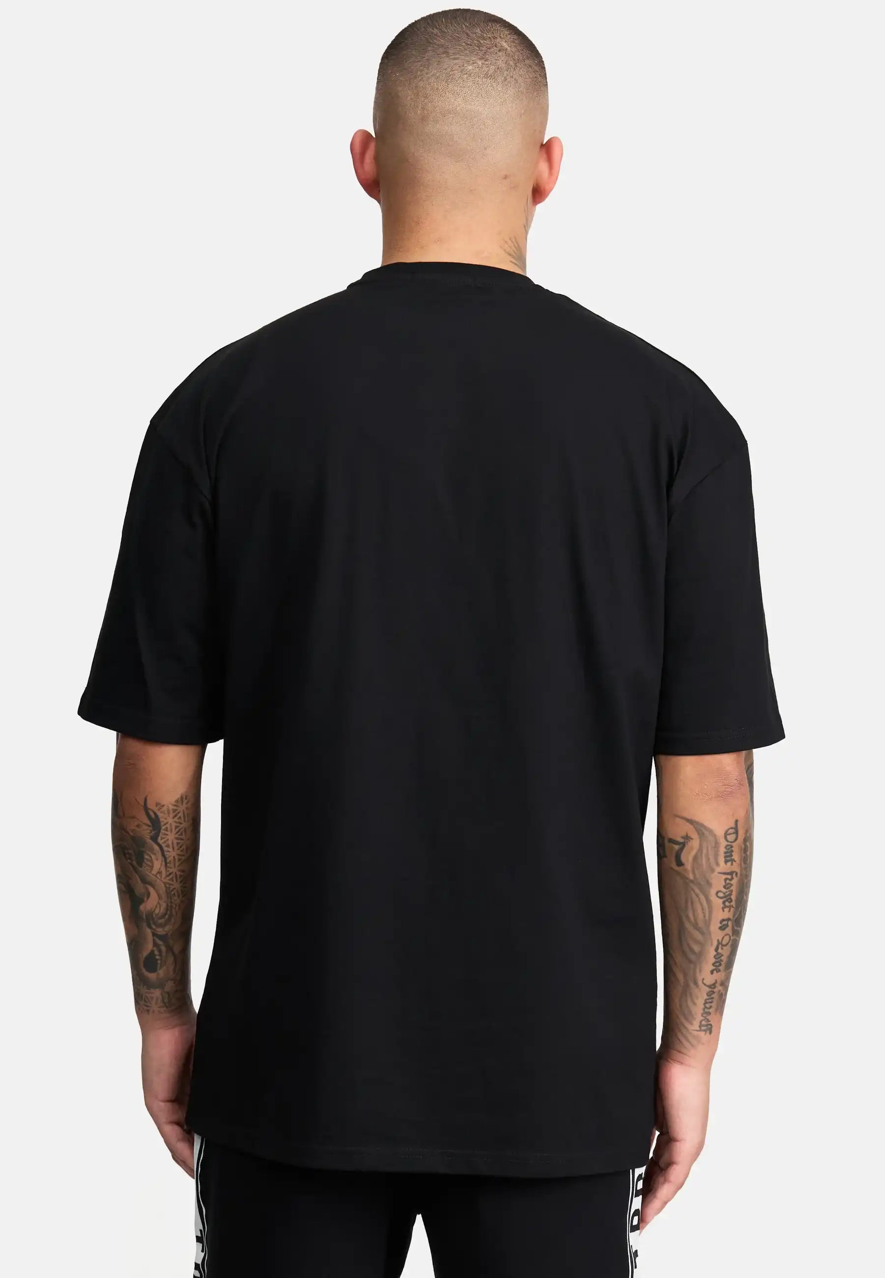 Gezeigt wird eine tätowierte Person mit kurzen Haaren von hinten, die das trueprodigy Miles Oversized T-Shirt mit Druck in schwarz und eine schwarze Hose trägt. Der Hintergrund ist einfarbig weiß.