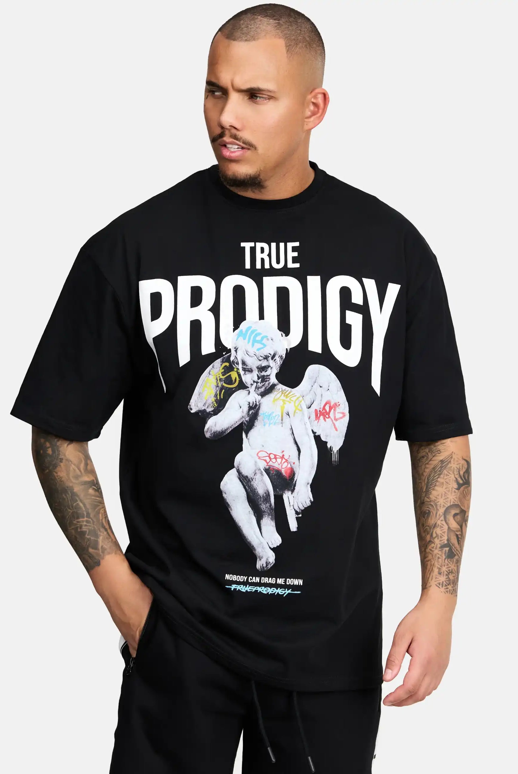Ein Mann mit kurzen Haaren und Unterarmtattoos trägt das trueprodigy Miles Oversized T-Shirt mit Druck, das einen fetten weißen TRUE PRODIGY-Schriftzug und eine mit Graffiti überzogene steinerne Engelsgrafik vor einem schlichten hellen Hintergrund zeigt.
