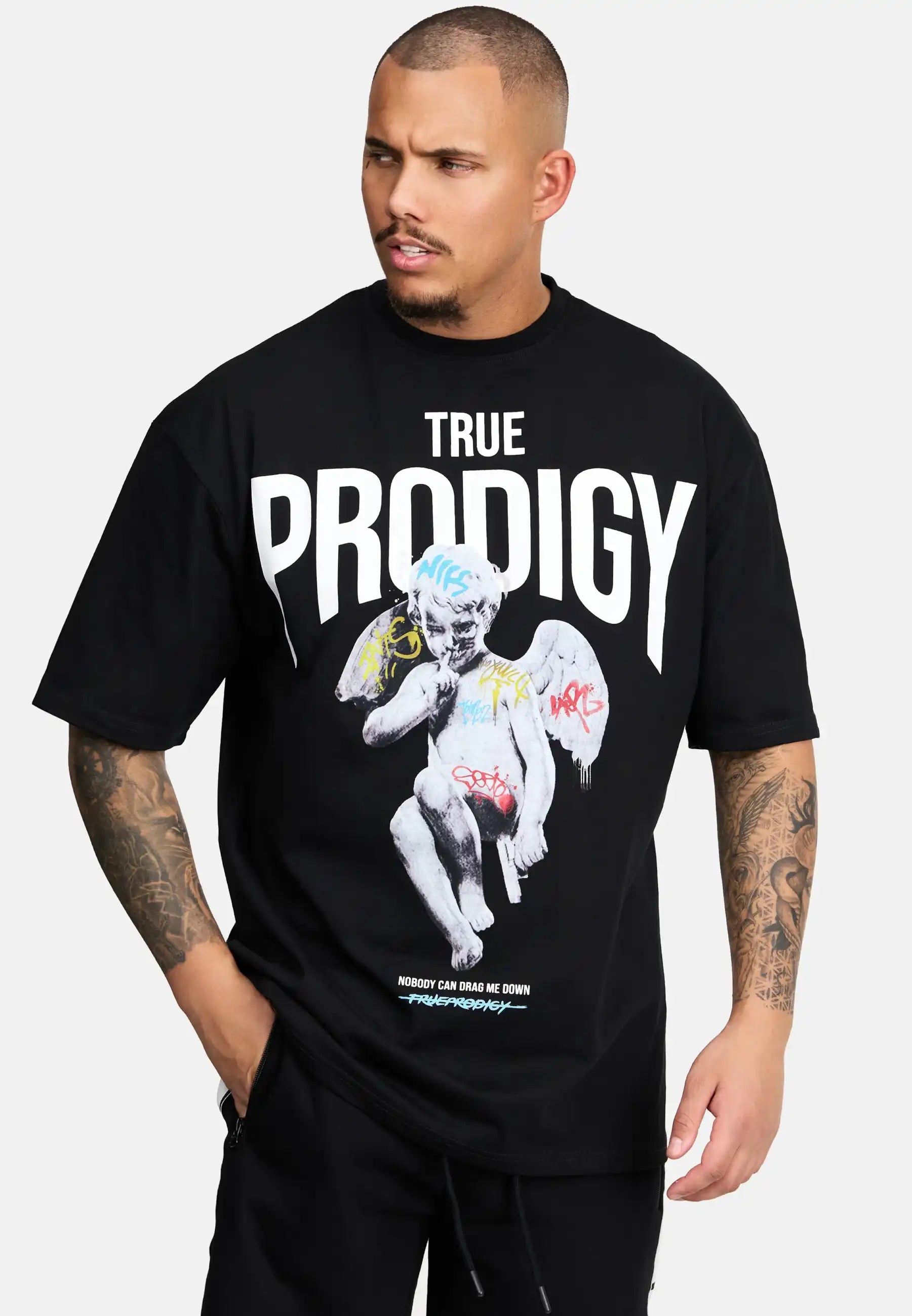 Ein Mann mit kurzen Haaren und Unterarmtattoos trägt das trueprodigy Miles Oversized T-Shirt mit Druck, das einen fetten weißen TRUE PRODIGY-Schriftzug und eine mit Graffiti überzogene steinerne Engelsgrafik vor einem schlichten hellen Hintergrund zeigt.