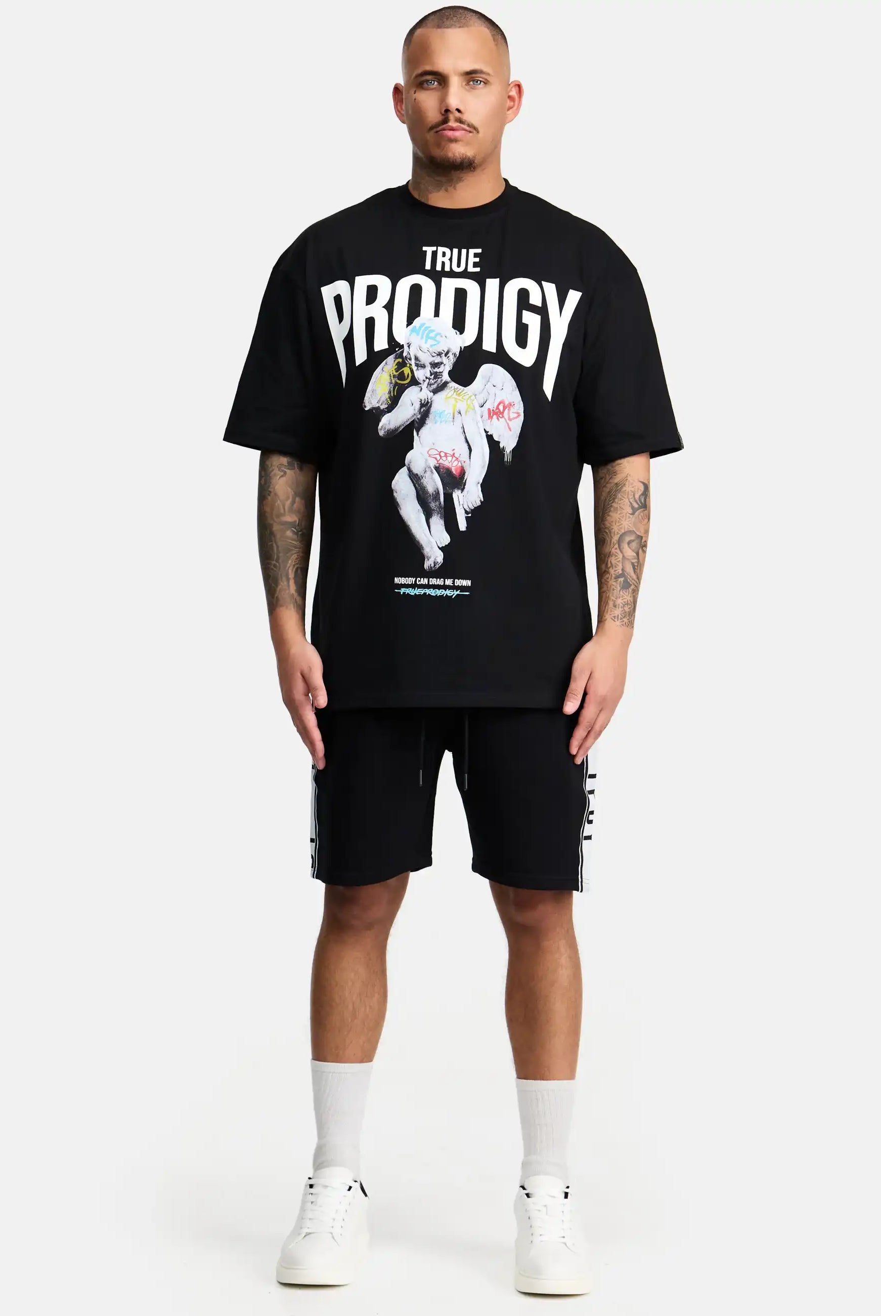 Ein Mann blickt nach vorne und trägt das trueprodigy Miles Oversized T-Shirt mit Druck in Schwarz, mit Text und einem grafischen Druck. Er kombiniert es mit schwarzen Shorts, weißen Socken und Sneakers. Der schwere Jersey-Stoff kontrastiert mit dem schlichten weißen Hintergrund.