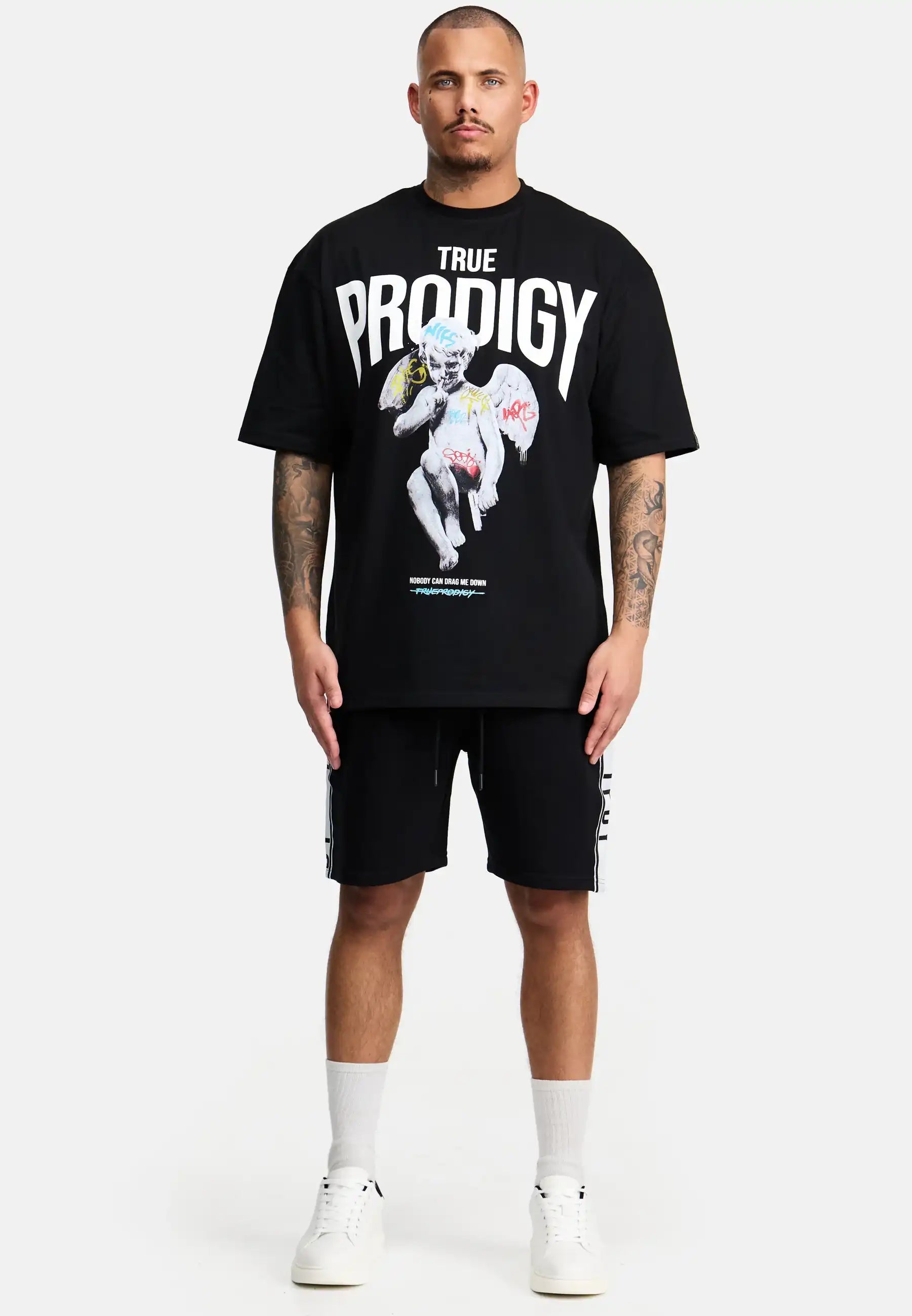 Ein Mann blickt nach vorne und trägt das trueprodigy Miles Oversized T-Shirt mit Druck in Schwarz, mit Text und einem grafischen Druck. Er kombiniert es mit schwarzen Shorts, weißen Socken und Sneakers. Der schwere Jersey-Stoff kontrastiert mit dem schlichten weißen Hintergrund.