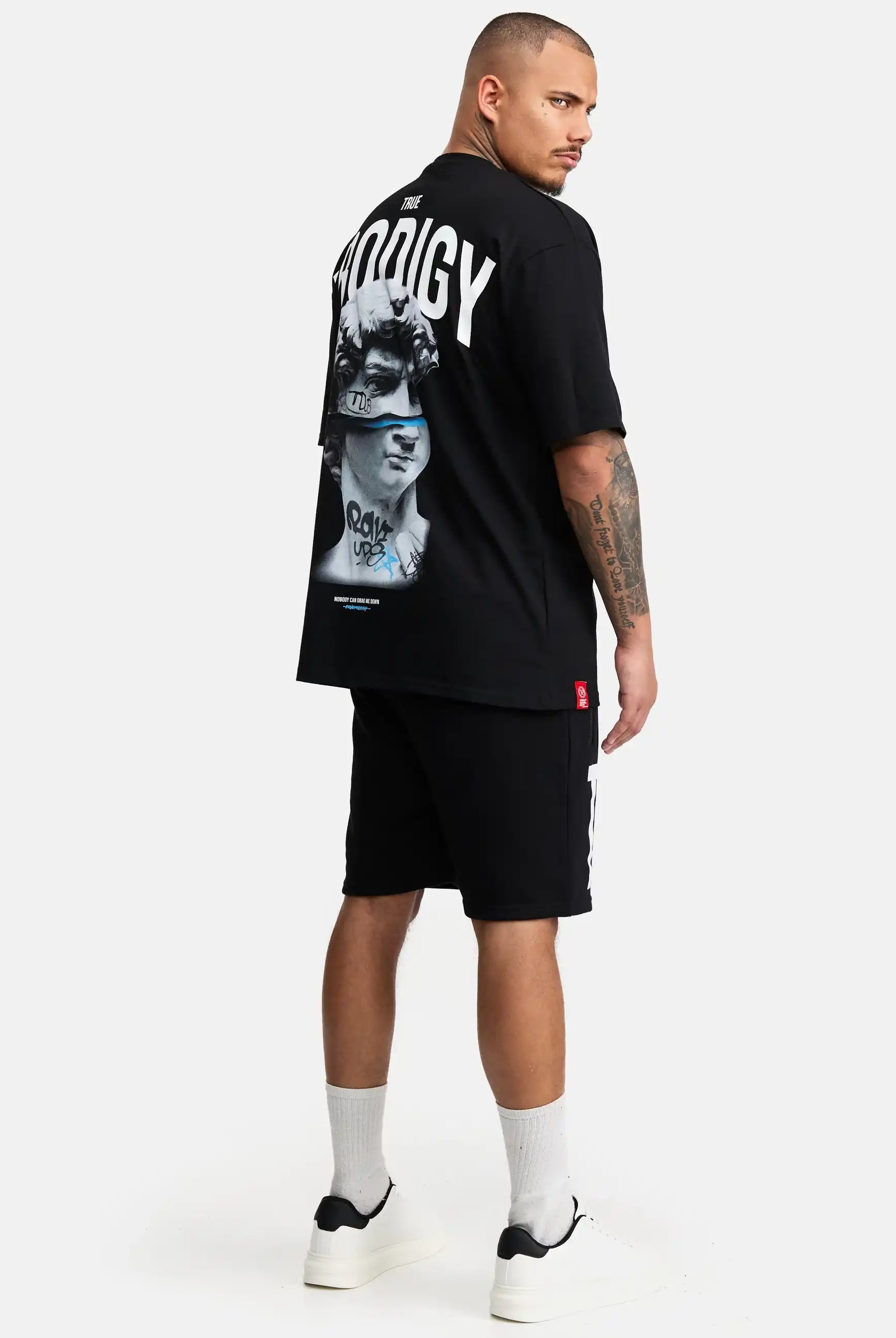 Ein Mann steht in einem schwarzen trueprodigy Bengi Oversized T-Shirt mit Druck mit "PRODIGY" auf dem Rücken, gepaart mit schwarzen Shorts, weißen Socken und schwarz-weißen Turnschuhen vor einem schlichten weißen Hintergrund.