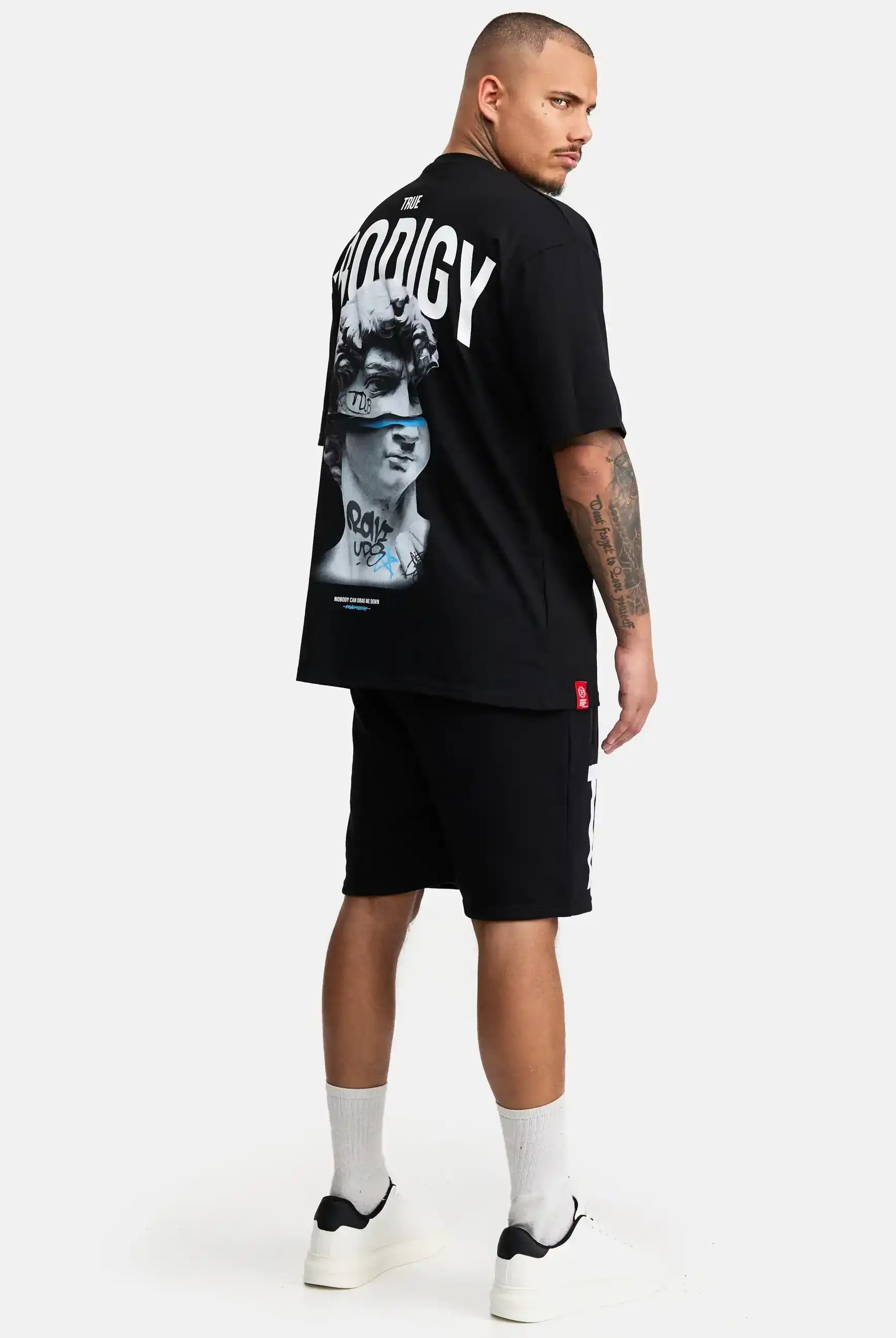 Ein Mann steht in einem schwarzen trueprodigy Bengi Oversized T-Shirt mit Druck mit "PRODIGY" auf dem Rücken, gepaart mit schwarzen Shorts, weißen Socken und schwarz-weißen Turnschuhen vor einem schlichten weißen Hintergrund.
