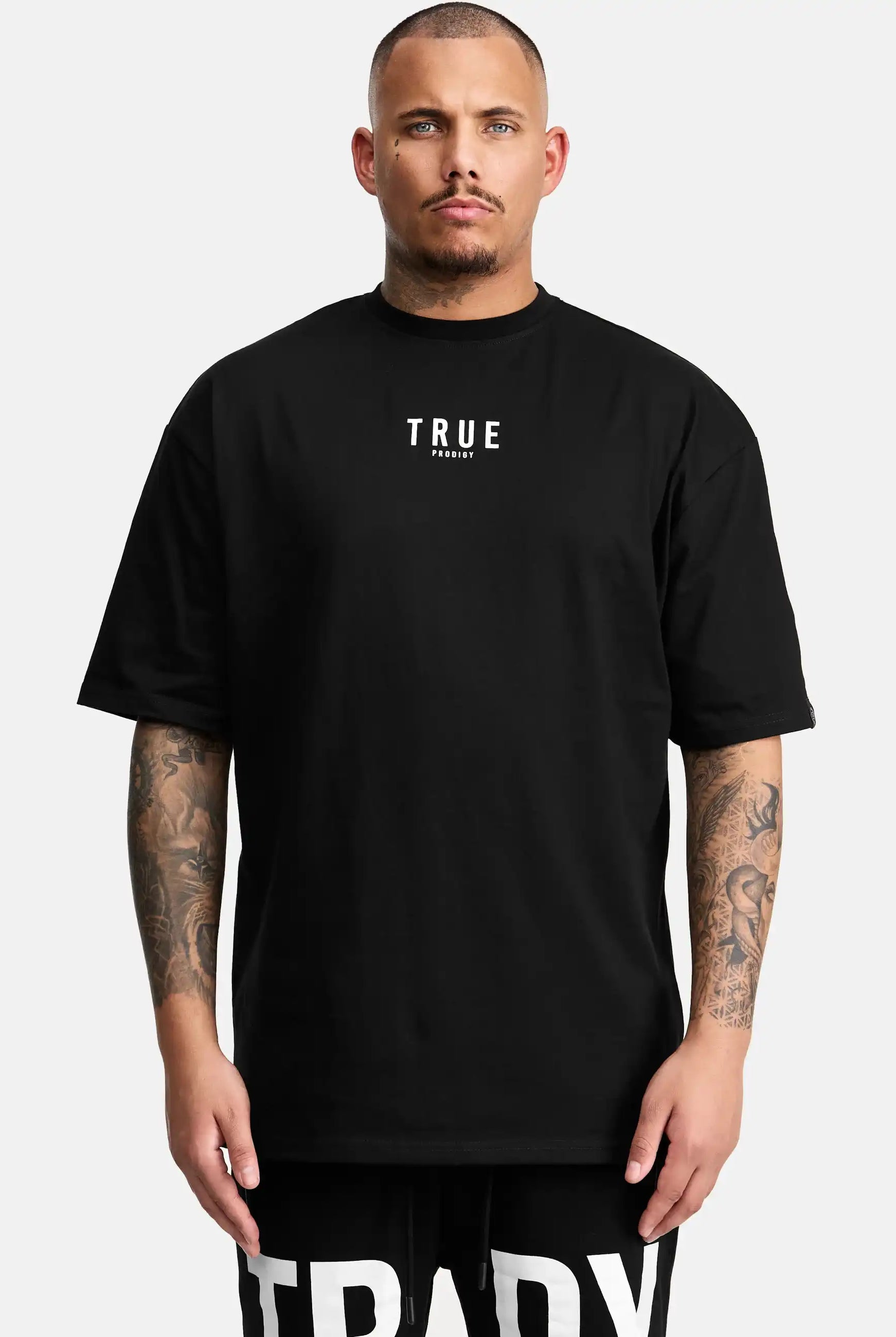 Eine Person steht mit dem Gesicht nach vorne, trägt das trueprodigy Bengi Oversized T-Shirt mit Druck in schwarz mit "TRUE" in weiß auf der Brust und schwarzen Shorts, hebt Tattoos auf beiden Armen und einen neutralen Ausdruck hervor.