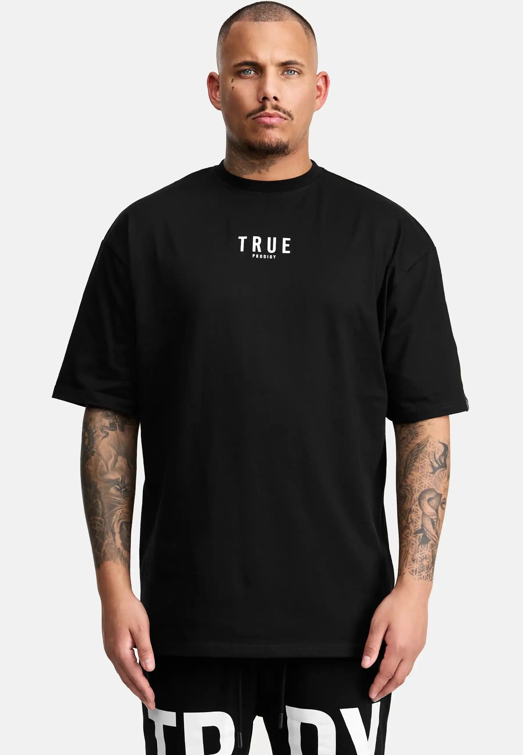Eine Person steht mit dem Gesicht nach vorne, trägt das trueprodigy Bengi Oversized T-Shirt mit Druck in schwarz mit "TRUE" in weiß auf der Brust und schwarzen Shorts, hebt Tattoos auf beiden Armen und einen neutralen Ausdruck hervor.