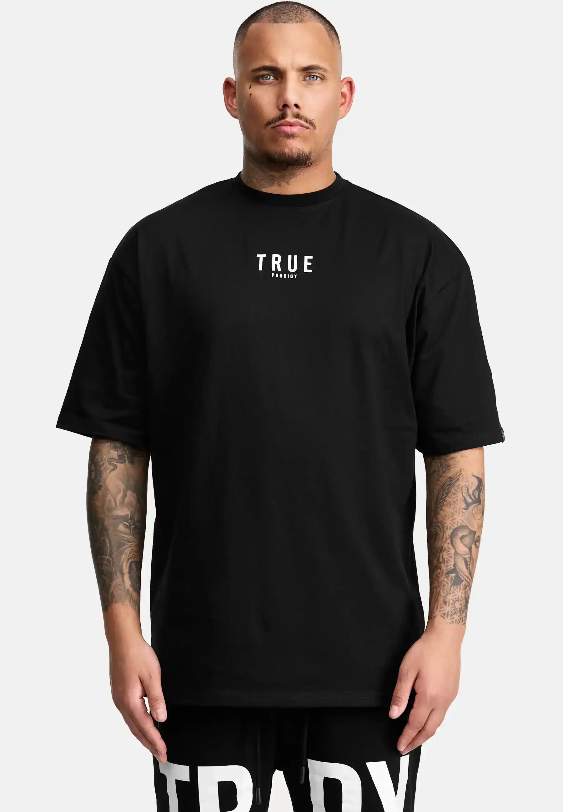 Eine Person steht mit dem Gesicht nach vorne, trägt das trueprodigy Bengi Oversized T-Shirt mit Druck in schwarz mit "TRUE" in weiß auf der Brust und schwarzen Shorts, hebt Tattoos auf beiden Armen und einen neutralen Ausdruck hervor.
