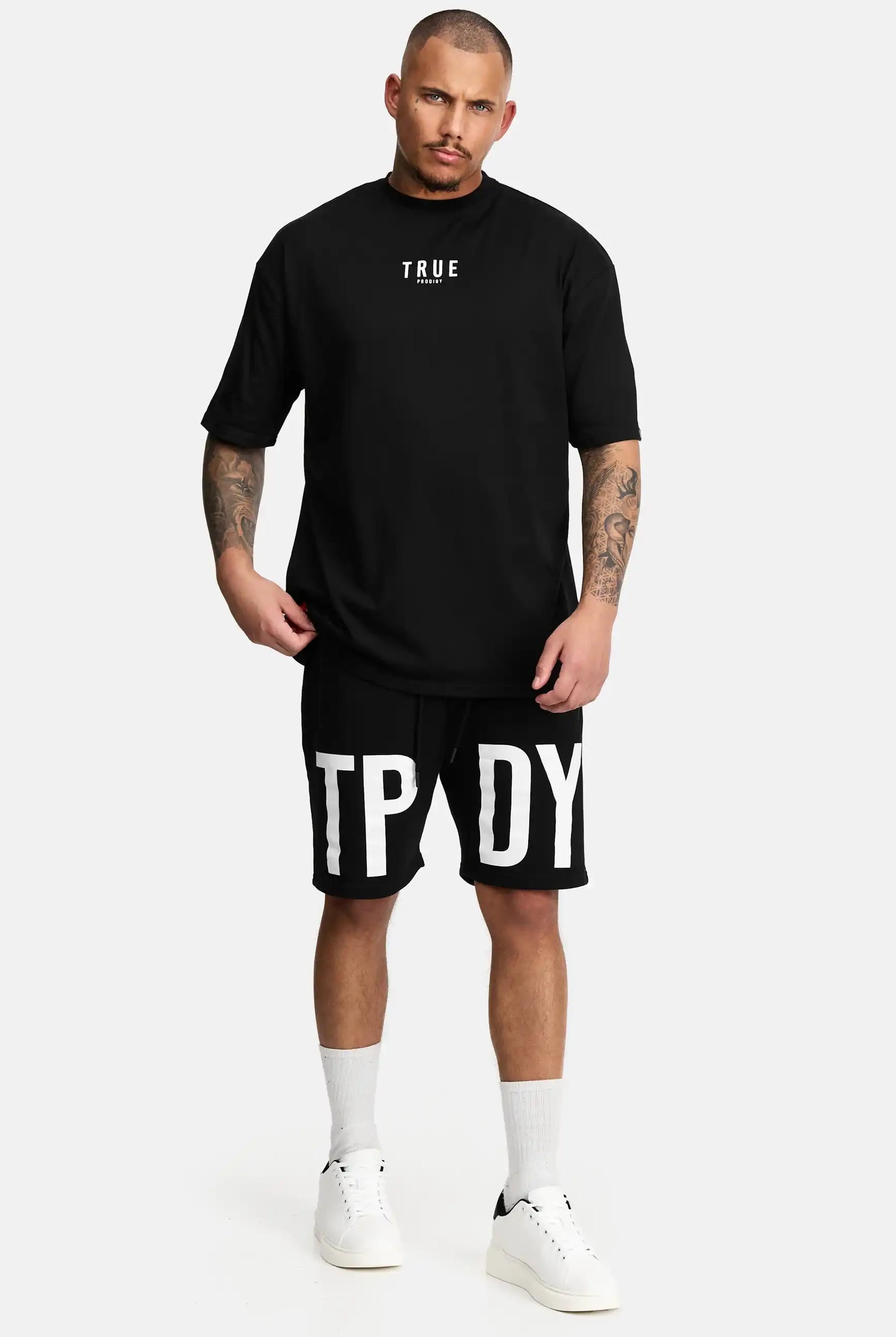 Ein Mann schaut nach vorne und trägt das trueprodigy Bengi Oversized T-Shirt mit Druck in schwarz, gepaart mit schwarzen Shorts mit großen weißen Buchstaben, weißen Socken und weißen Turnschuhen vor einem schlichten, hellen Hintergrund.