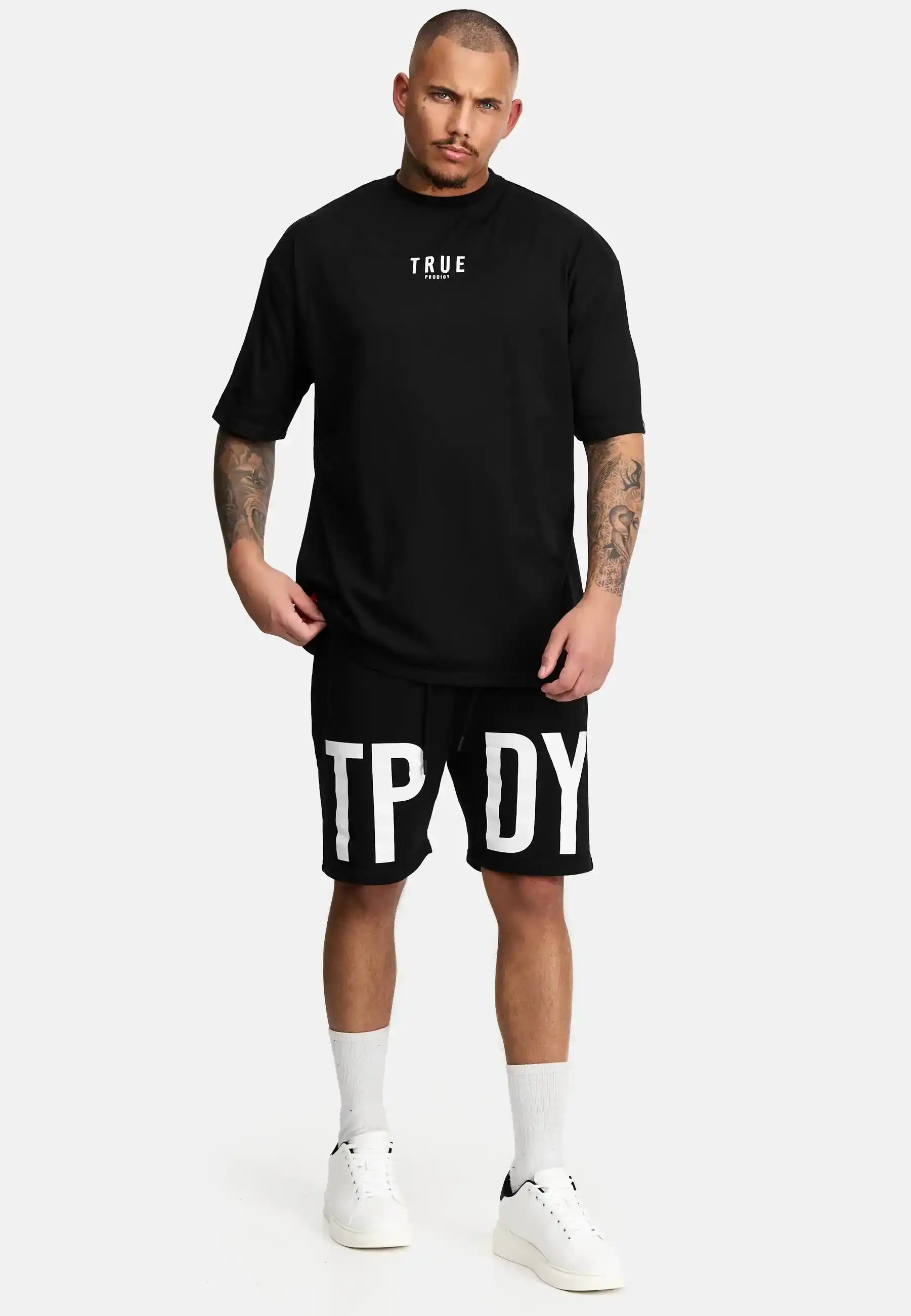 Ein Mann schaut nach vorne und trägt das trueprodigy Bengi Oversized T-Shirt mit Druck in schwarz, gepaart mit schwarzen Shorts mit großen weißen Buchstaben, weißen Socken und weißen Turnschuhen vor einem schlichten, hellen Hintergrund.