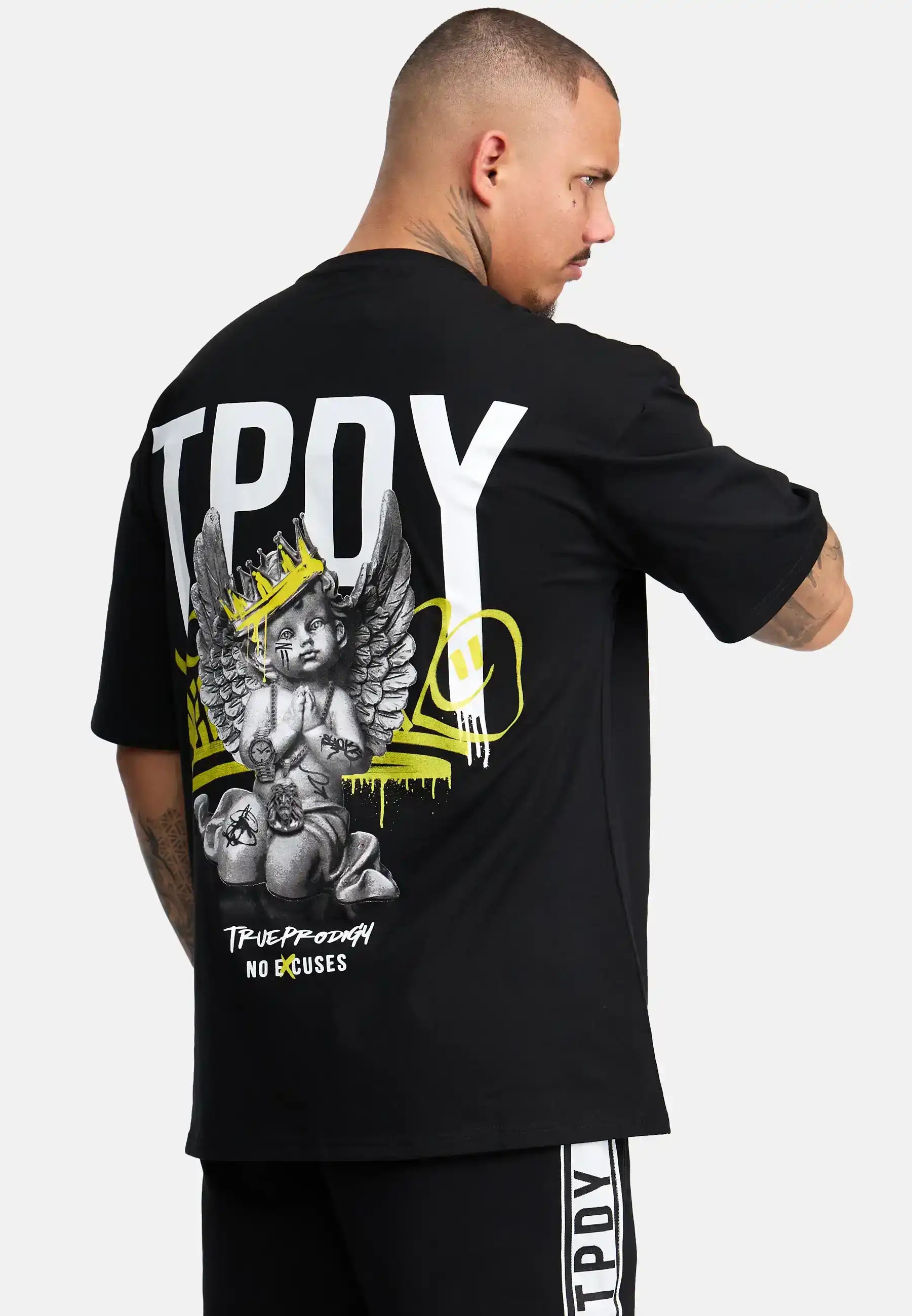 Ein Mann mit kurzen Haaren und Tattoos trägt das trueprodigy Jinx Oversized T-Shirt mit Druck: ein schwarzes Unisex-T-Shirt aus schwerem Jersey mit "TPOY" in großen weißen Buchstaben, einem Engel, der einen Totenkopf hält, und "TRUE PRODIGY NO EXCUSES" am Saum.
