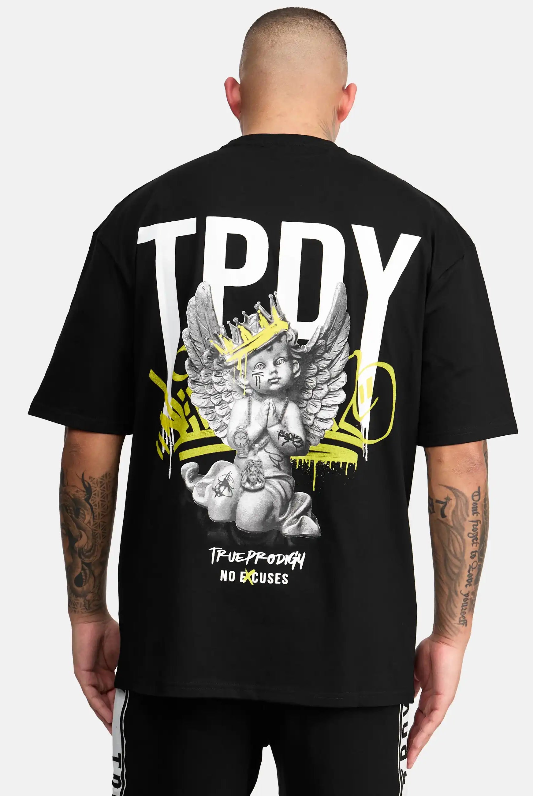 Eine Person blickt weg und trägt das trueprodigy Jinx Oversized T-Shirt in Schwarz. Auf der Rückseite befindet sich ein großer weißer "TPDY"-Schriftzug, ein Engel mit einer Krone, gelbe Graffiti-Akzente und "TRUE PRODIGY NO EXCUSES" am unteren Rand.