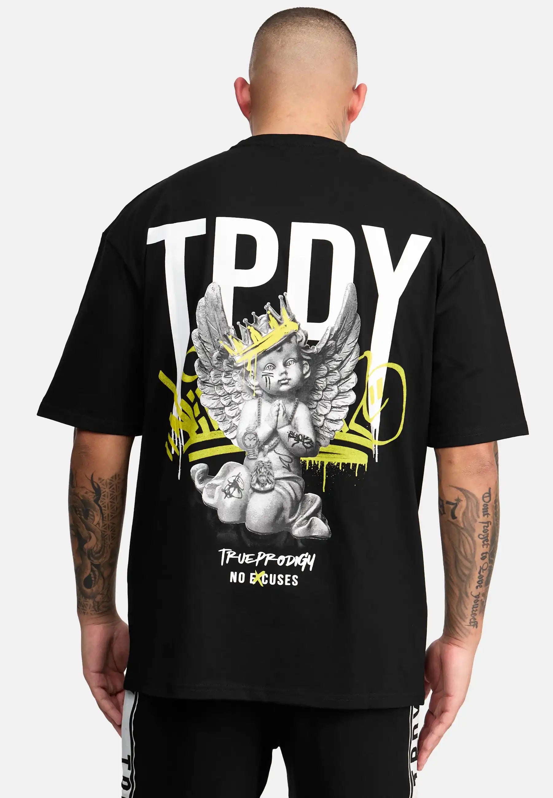 Eine Person blickt weg und trägt das trueprodigy Jinx Oversized T-Shirt in Schwarz. Auf der Rückseite befindet sich ein großer weißer "TPDY"-Schriftzug, ein Engel mit einer Krone, gelbe Graffiti-Akzente und "TRUE PRODIGY NO EXCUSES" am unteren Rand.