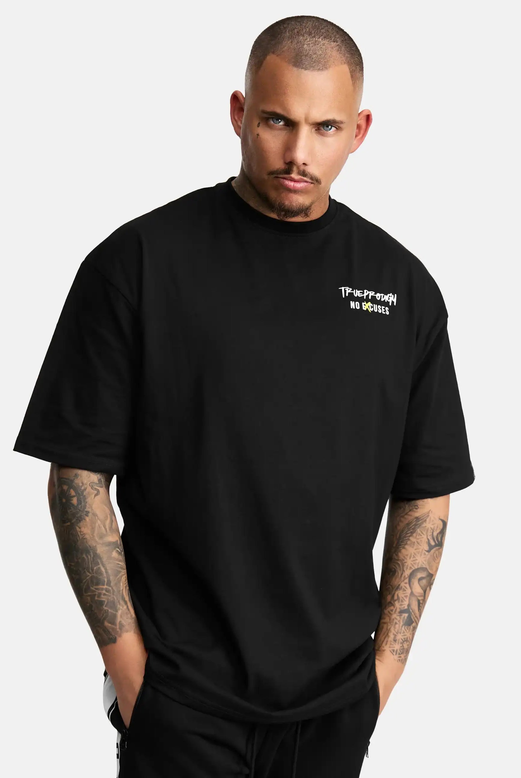 Ein Mann mit rasierter Frisur und Armtattoos trägt das Jinx Oversized T-Shirt mit Druck von trueprodigy, ein Unisex-T-Shirt aus schwerem Jersey mit dem Aufdruck "Takeoff Your No Brakes" in kleiner weißer Schrift auf der linken Brust, die Hände in den Taschen vor einem schlichten Hintergrund.