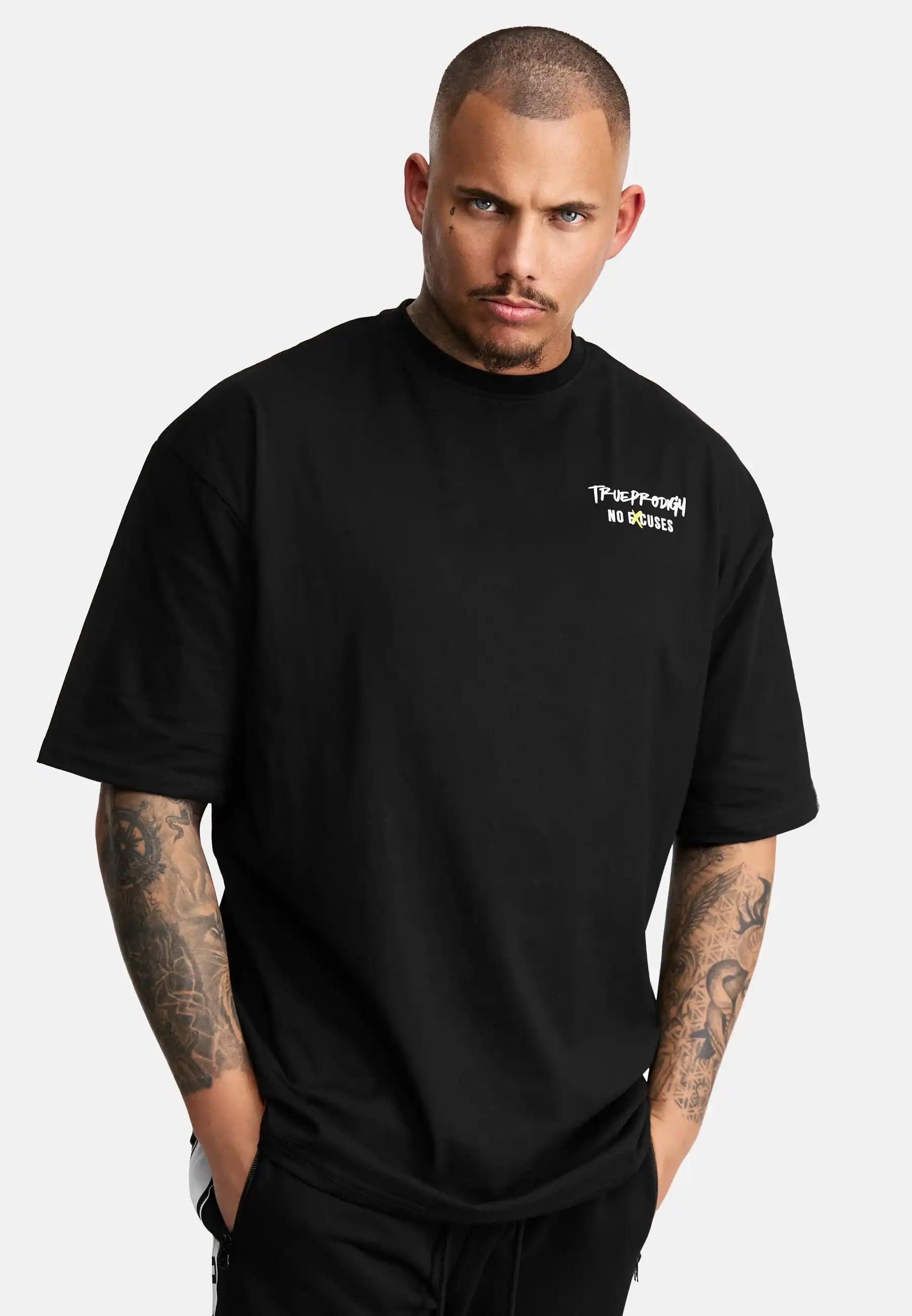 Ein Mann mit rasierter Frisur und Armtattoos trägt das Jinx Oversized T-Shirt mit Druck von trueprodigy, ein Unisex-T-Shirt aus schwerem Jersey mit dem Aufdruck "Takeoff Your No Brakes" in kleiner weißer Schrift auf der linken Brust, die Hände in den Taschen vor einem schlichten Hintergrund.