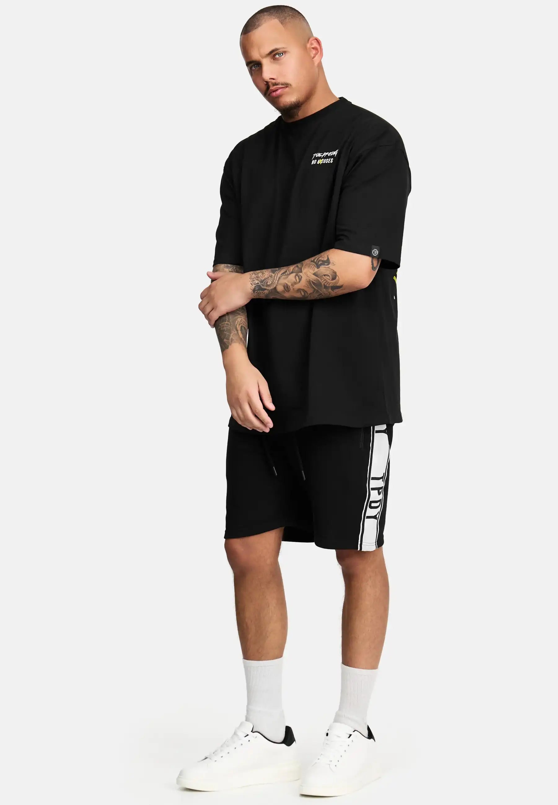 Ein Mann mit kurzen Haaren und gestutztem Bart steht vor einem schlichten Hintergrund und trägt das schwarze trueprodigy Jinx Oversized T-Shirt mit Druck, schwarze Shorts mit weißen Seitenstreifen, weiße Socken und weiße Turnschuhe.