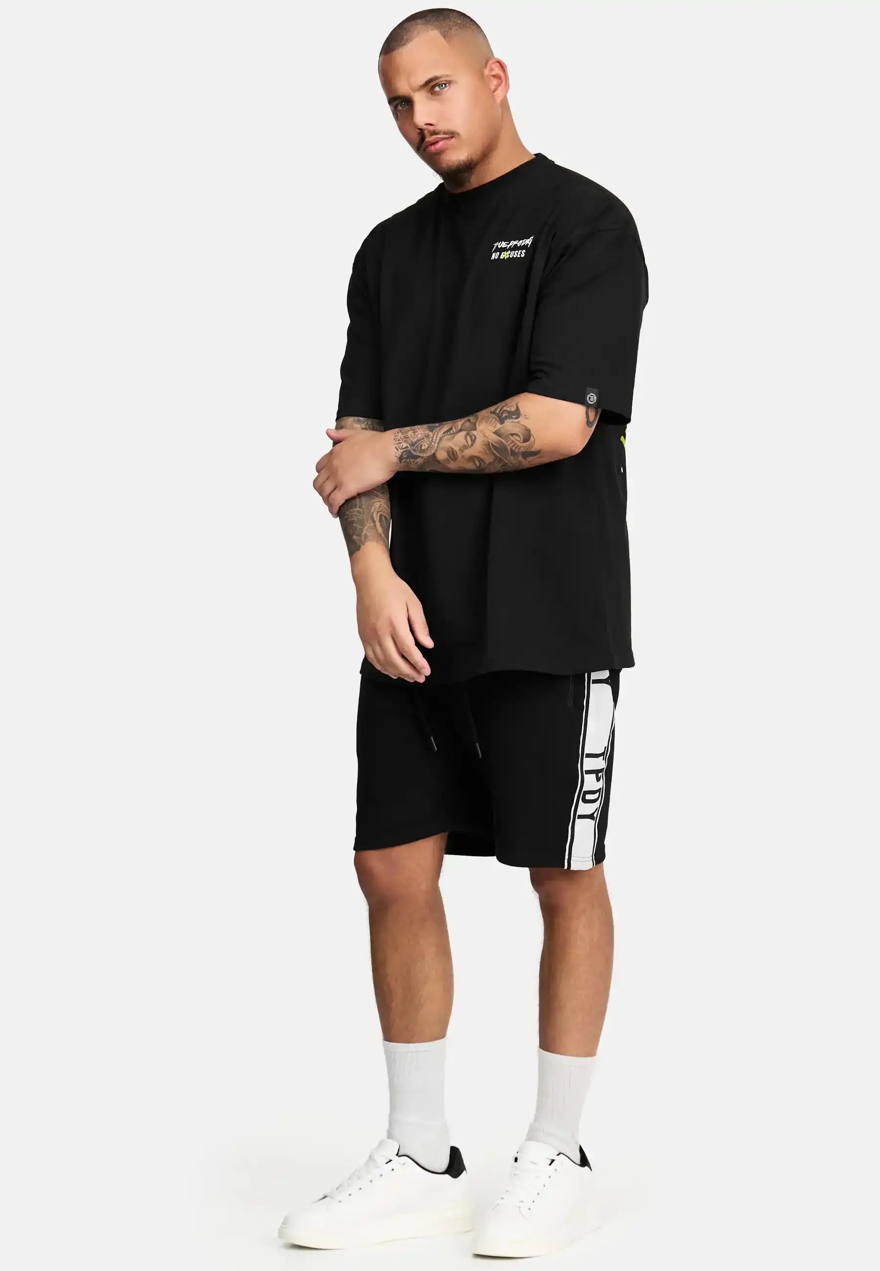 Ein Mann mit kurzen Haaren und gestutztem Bart steht vor einem schlichten Hintergrund und trägt das schwarze trueprodigy Jinx Oversized T-Shirt mit Druck, schwarze Shorts mit weißen Seitenstreifen, weiße Socken und weiße Turnschuhe.