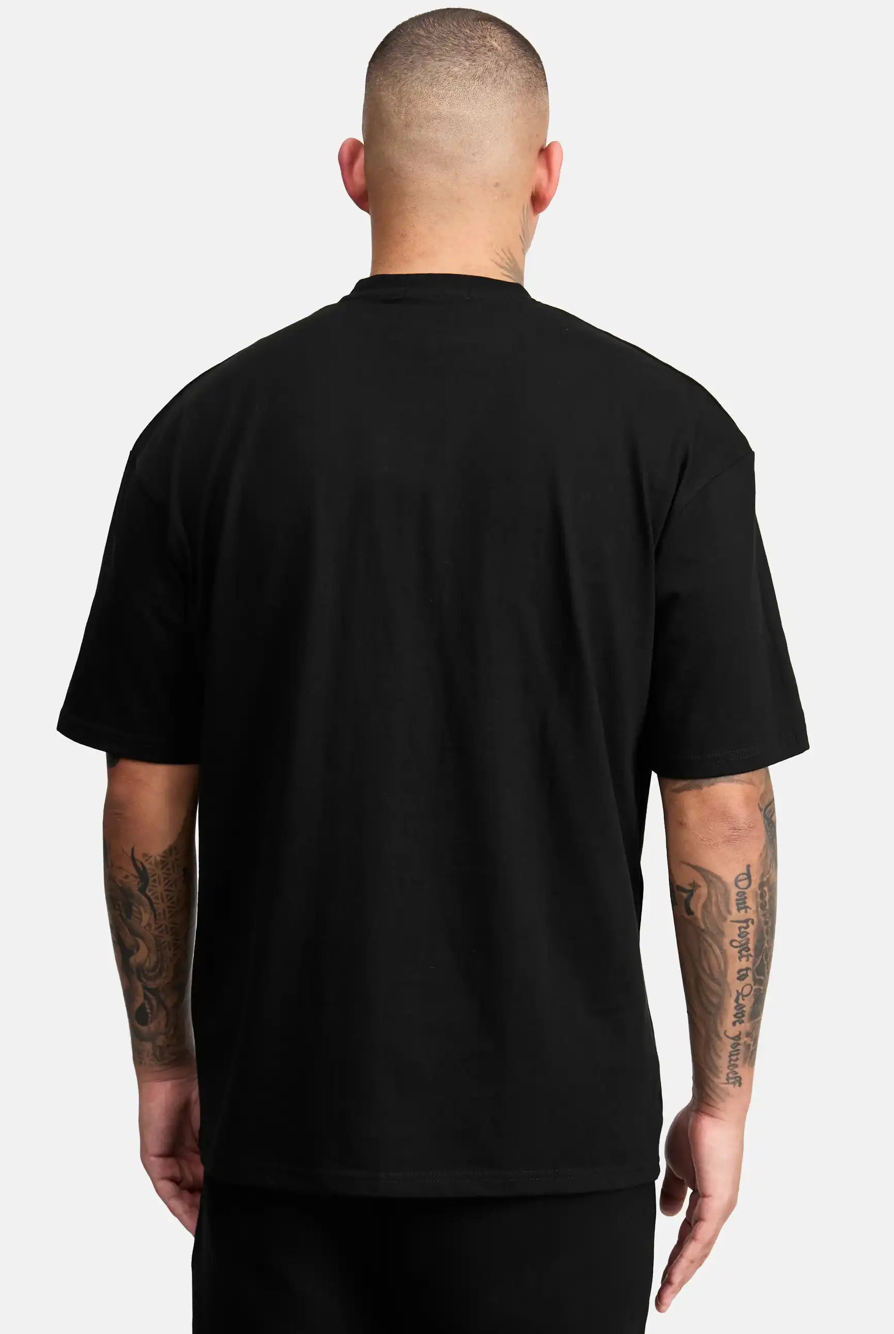 Eine Person mit kurzen Haaren und tätowierten Armen steht mit dem Rücken zur Kamera, trägt das trueprodigy Dexter Oversized T-Shirt mit Druck und eine schwarze Hose vor einem schlichten, hellen Hintergrund.