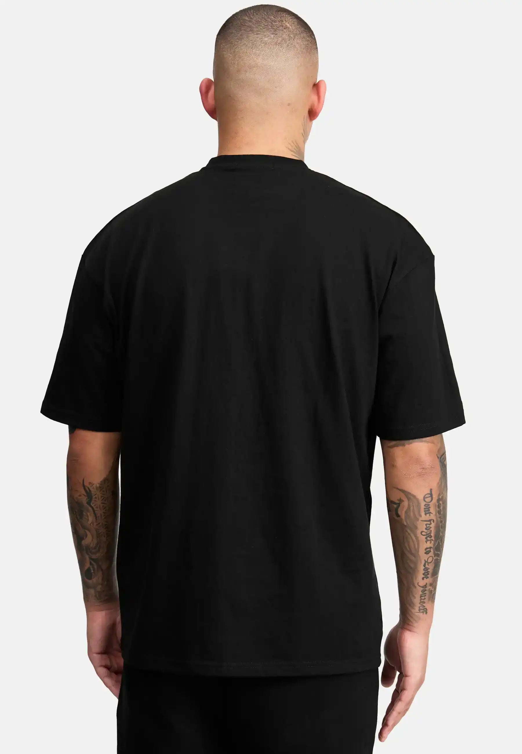 Eine Person mit kurzen Haaren und tätowierten Armen steht mit dem Rücken zur Kamera, trägt das trueprodigy Dexter Oversized T-Shirt mit Druck und eine schwarze Hose vor einem schlichten, hellen Hintergrund.