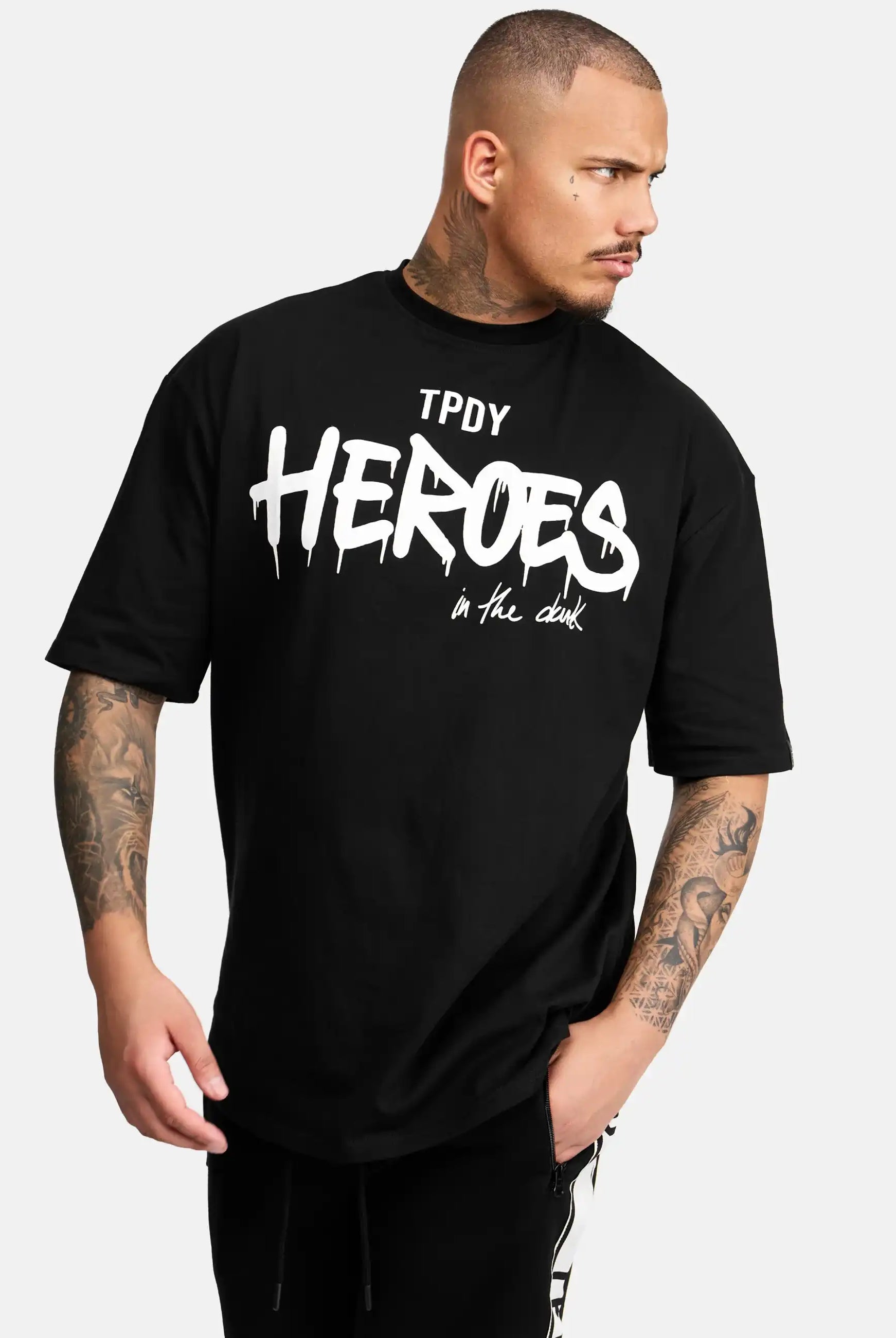 Eine Person mit kurzen Haaren und Tattoos auf beiden Armen trägt das trueprodigy Dexter Oversized T-Shirt mit Druck in schwarz, mit weißem, stilisiertem "TPDY HEROES" Aufdruck. Sie schaut zur Seite und hat eine Hand lässig in der Tasche.