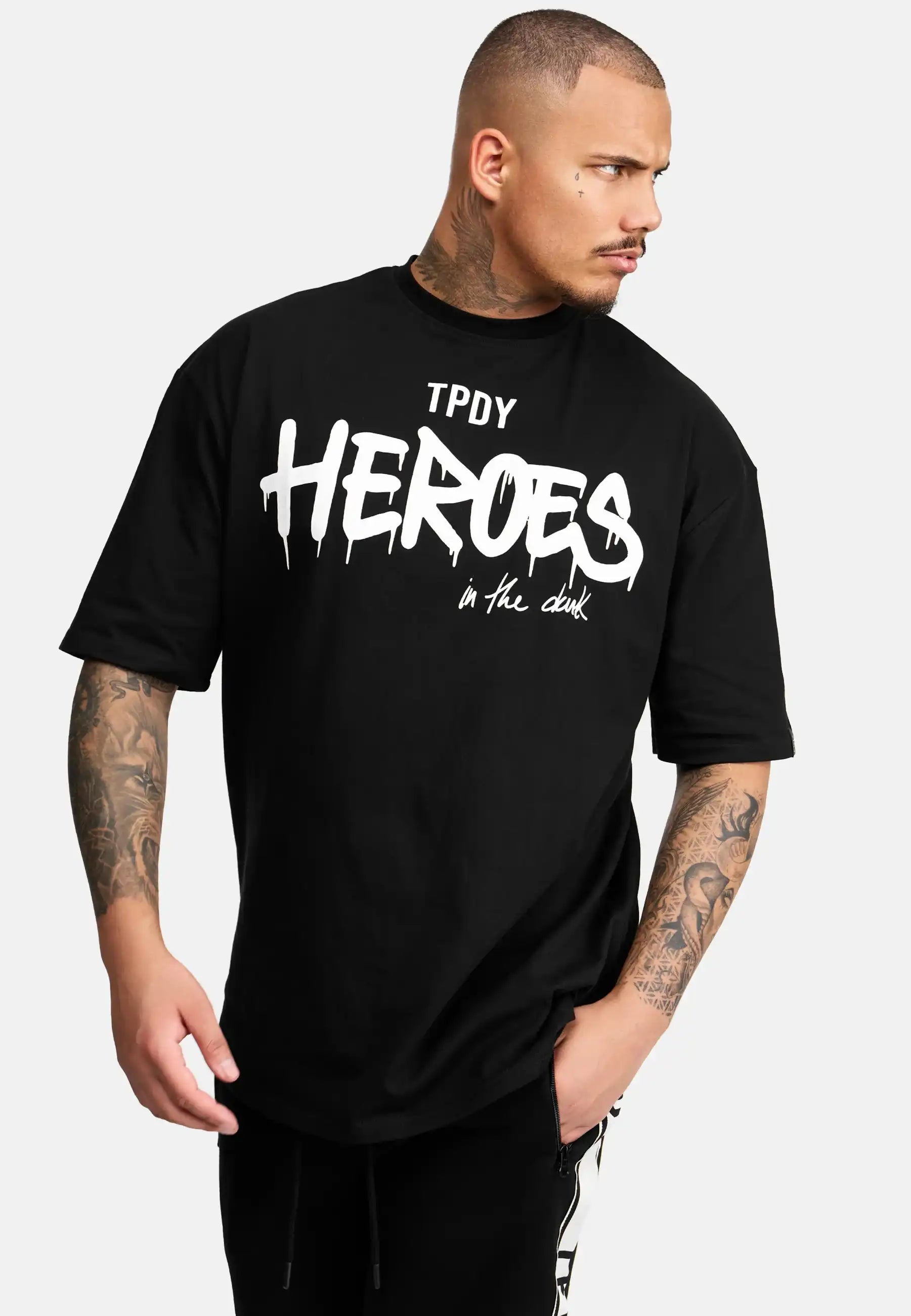 Eine Person mit kurzen Haaren und Tattoos auf beiden Armen trägt das trueprodigy Dexter Oversized T-Shirt mit Druck in schwarz, mit weißem, stilisiertem "TPDY HEROES" Aufdruck. Sie schaut zur Seite und hat eine Hand lässig in der Tasche.