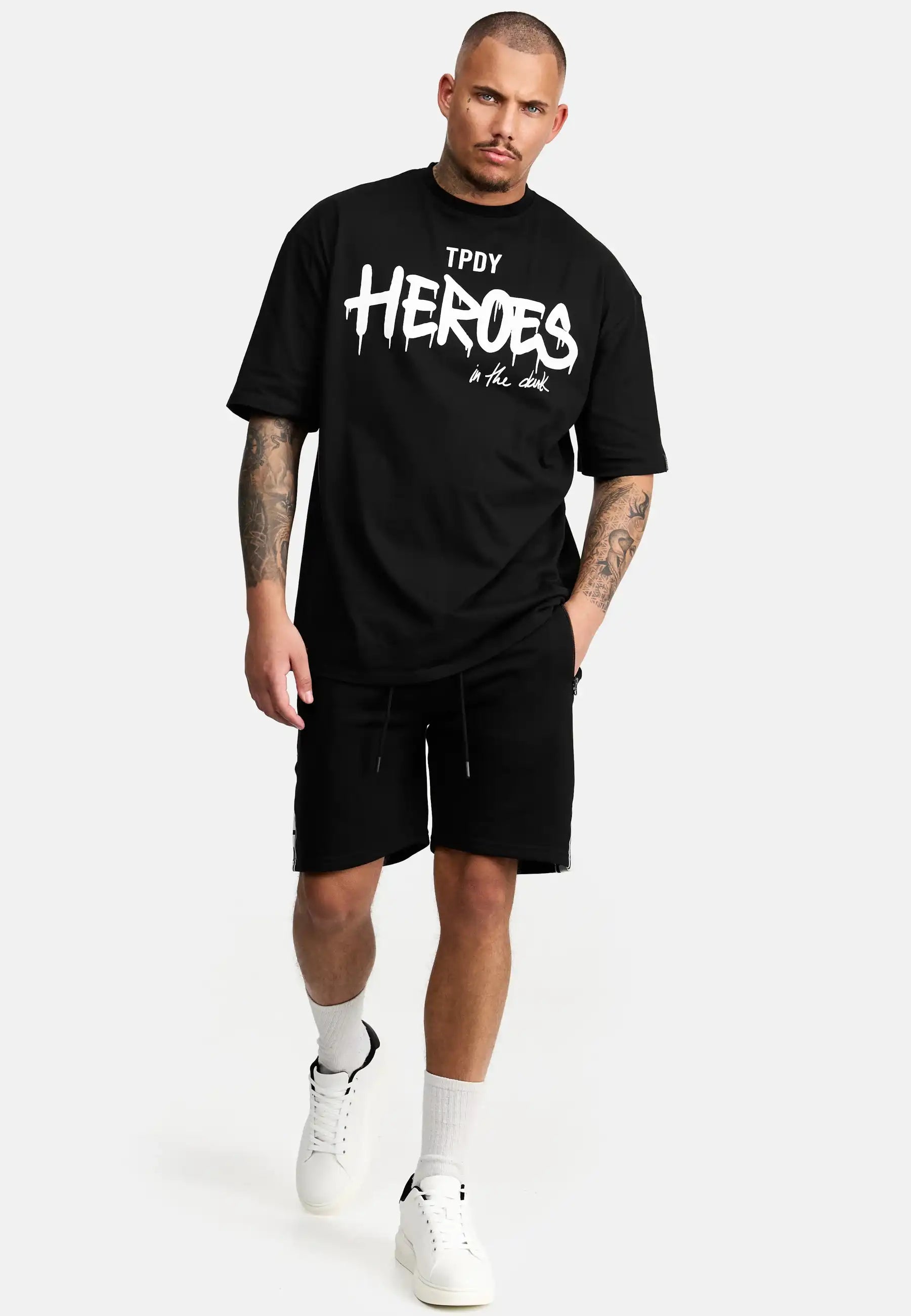 Ein Mann mit kurzen Haaren geht vor einem schlichten hellen Hintergrund nach vorne. Er trägt das trueprodigy Dexter Oversized T-Shirt mit Druck in schwarz, gepaart mit schwarzen Shorts, weißen Socken und weißen Turnschuhen.