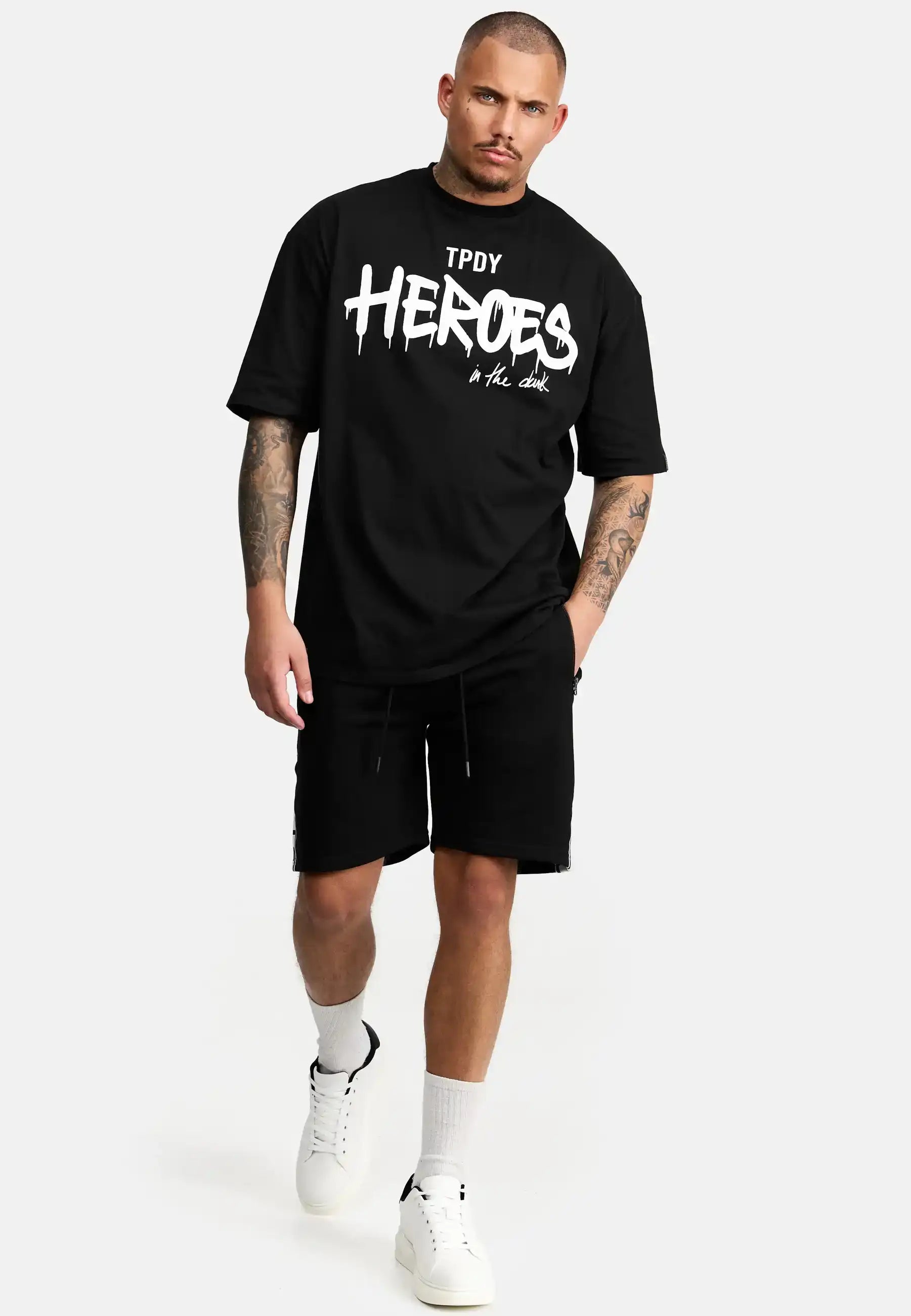 Ein Mann mit kurzen Haaren geht vor einem schlichten hellen Hintergrund nach vorne. Er trägt das trueprodigy Dexter Oversized T-Shirt mit Druck in schwarz, gepaart mit schwarzen Shorts, weißen Socken und weißen Turnschuhen.