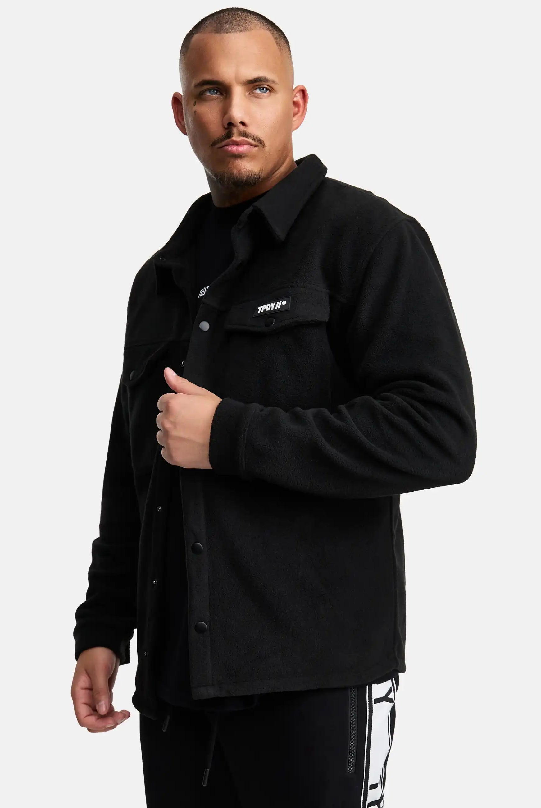 Ein Mann mit kurzen Haaren und gestutztem Bart trägt ein schwarzes Trueprodigy Bill Fleece Oversized Hemd über einem schwarzen Hemd mit weißer Aufschrift und einer weißen Hose mit schwarzen Streifen, stehend vor einem schlichten weißen Hintergrund.