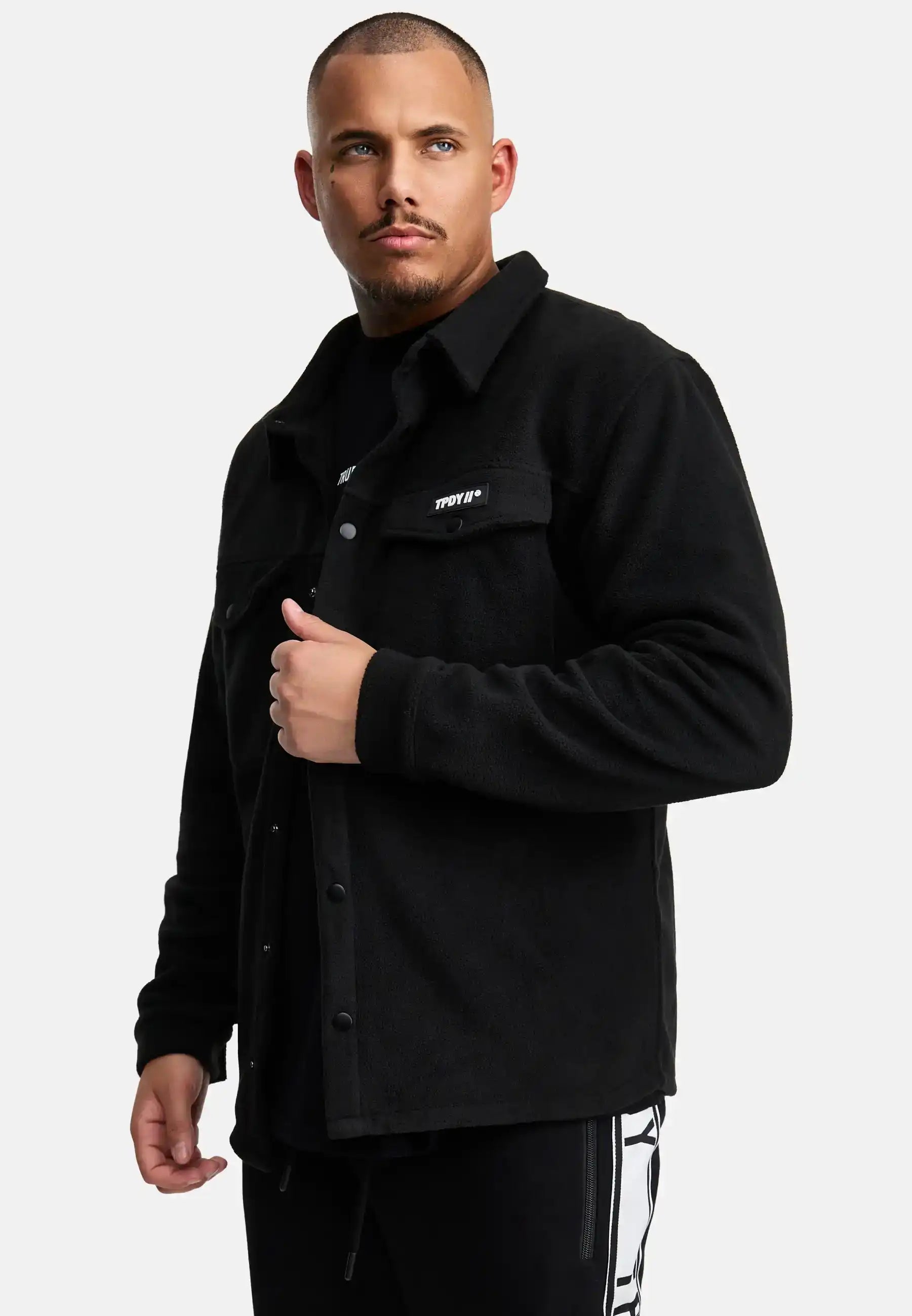 Ein Mann mit kurzen Haaren und gestutztem Bart trägt ein schwarzes Trueprodigy Bill Fleece Oversized Hemd über einem schwarzen Hemd mit weißer Aufschrift und einer weißen Hose mit schwarzen Streifen, stehend vor einem schlichten weißen Hintergrund.