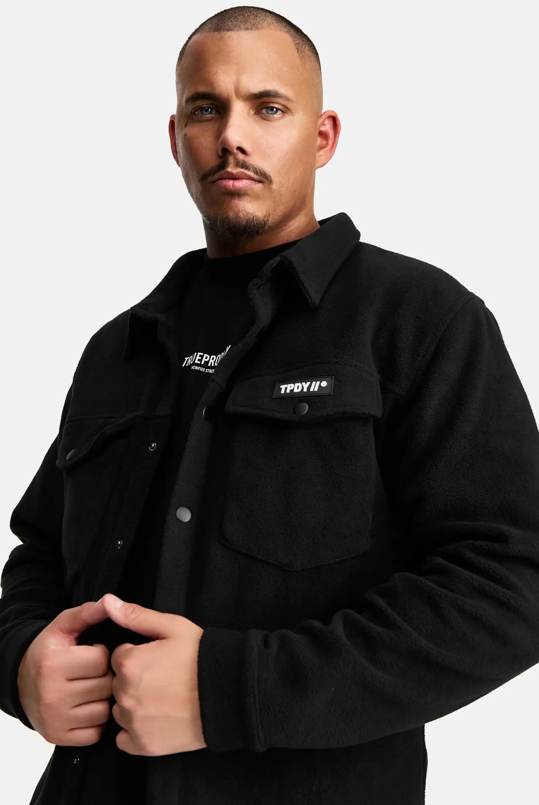 Ein Mann mit kurzgeschnittenem Haar und gestutztem Bart trägt das schwarze trueprodigy Bill Fleece Oversized Hemd mit Druckknöpfen und einer Brusttasche mit TPDYIN-Aufnäher über einem schwarzen Hemd auf weißem Grund.