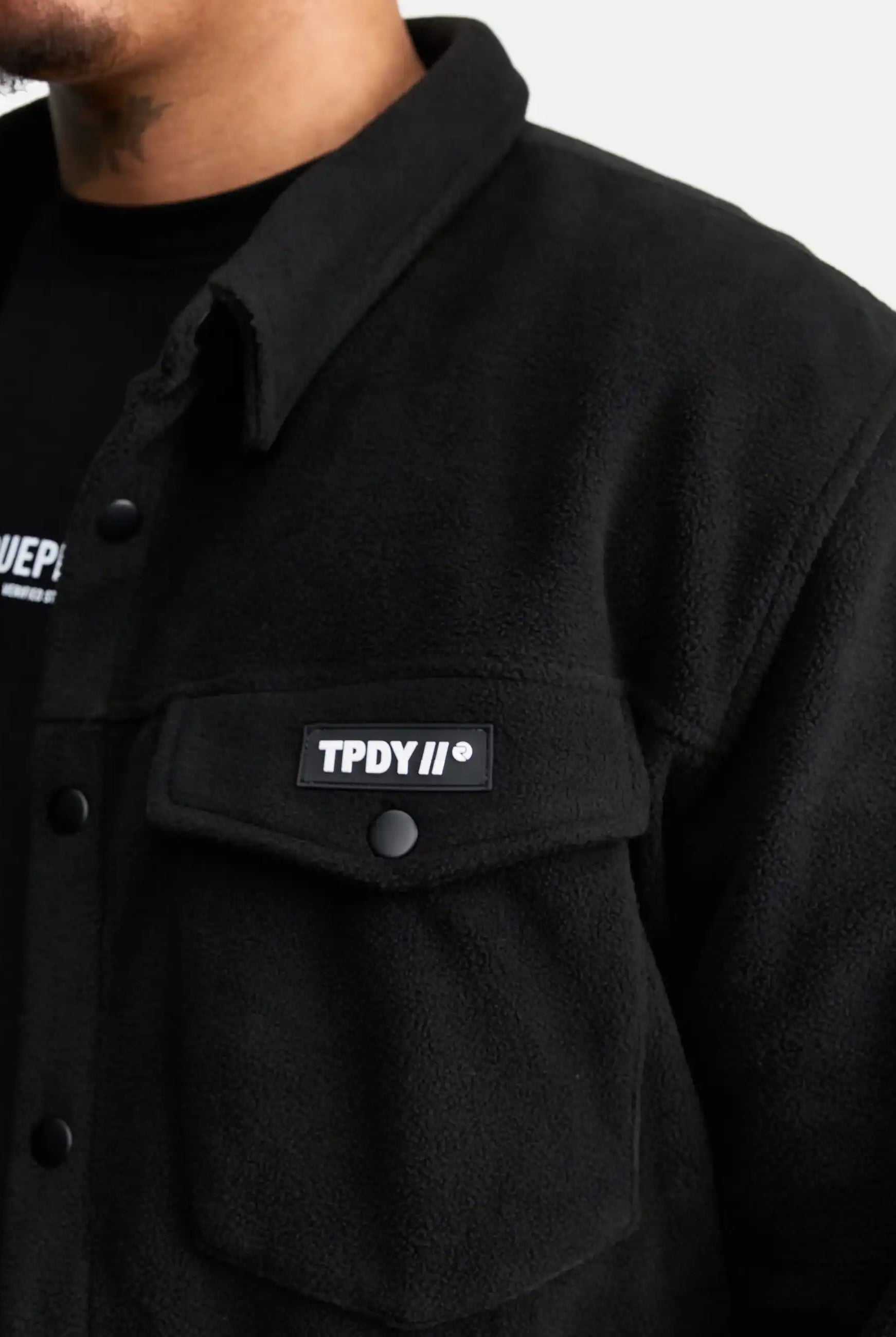 Eine Person trägt das trueprodigy Bill Fleece Oversized Hemd in Schwarz mit einer geknöpften Brusttasche mit einem rechteckigen TPDY II®-Aufnäher, der über einen weiteren übergroßen schwarzen Saum mit weißem Text gelegt ist.