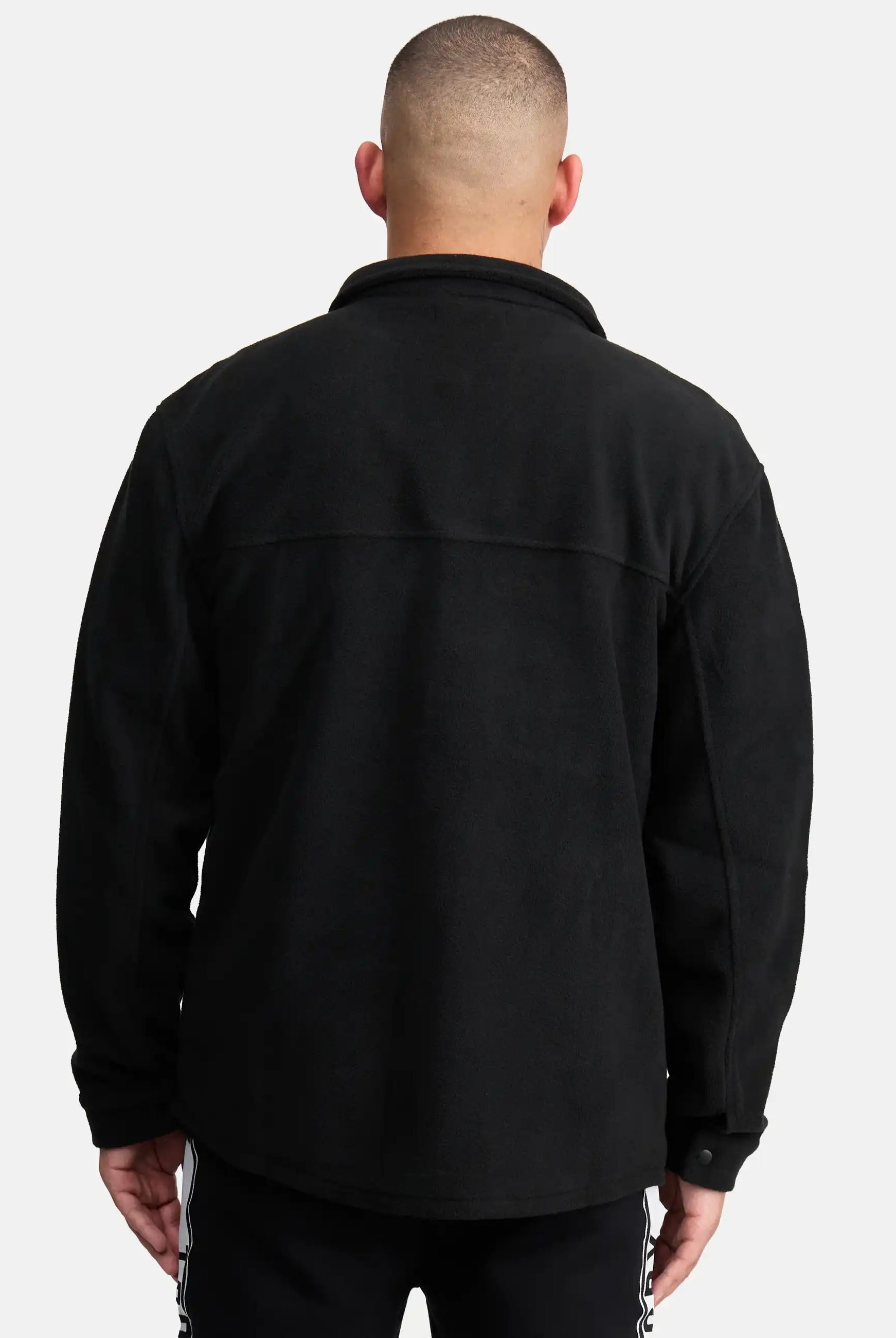 Ein Mann mit kurzen Haaren ist von hinten zu sehen und trägt ein schwarzes Trueprodigy Bill Fleece Oversized Hemd und eine schwarze Hose mit weißen Seitenstreifen. Er steht vor einem schlichten hellgrauen Hintergrund, der einen stilvollen, unaufdringlichen Look erzeugt.