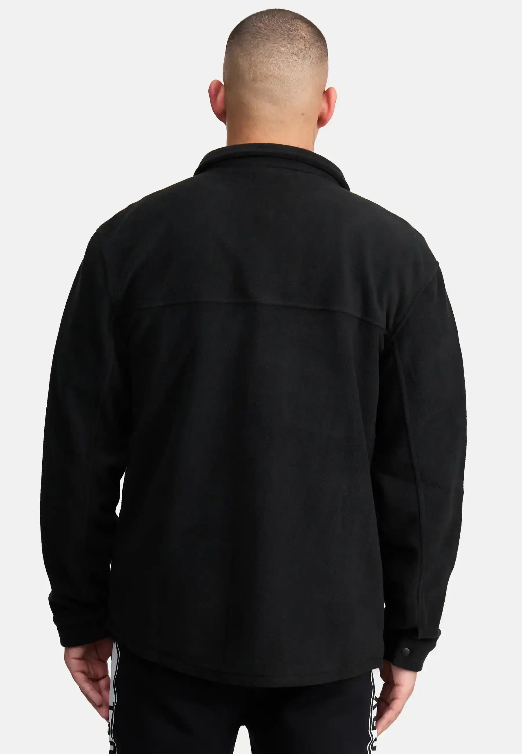 Ein Mann mit kurzen Haaren ist von hinten zu sehen und trägt ein schwarzes Trueprodigy Bill Fleece Oversized Hemd und eine schwarze Hose mit weißen Seitenstreifen. Er steht vor einem schlichten hellgrauen Hintergrund, der einen stilvollen, unaufdringlichen Look erzeugt.