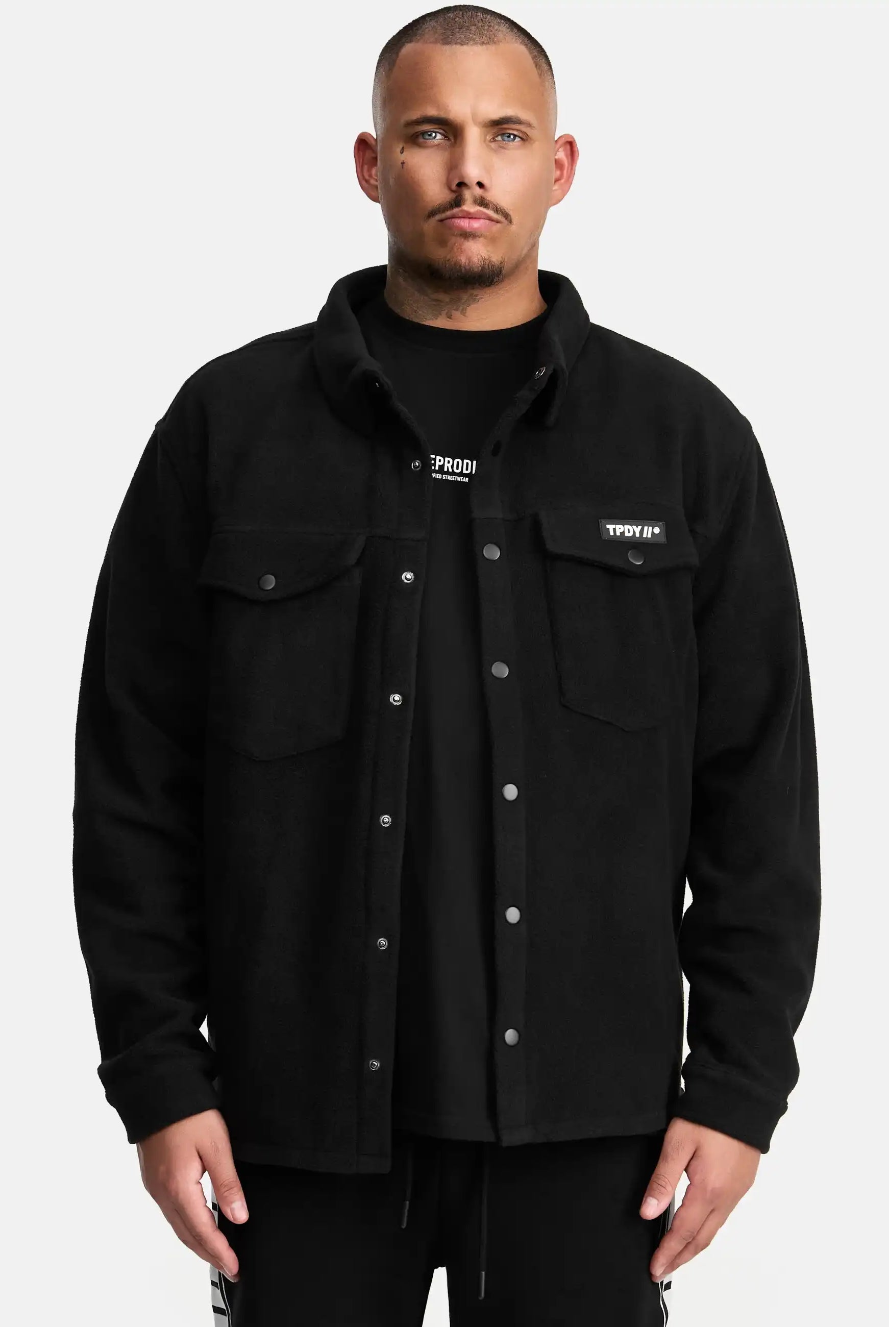Ein Mann blickt nach vorne in einem schwarzen Trueprodigy Bill Fleece Oversized Hemd über einem schwarzen T-Shirt. Das Hemd hat zwei Brusttaschen und ein kleines rechteckiges Etikett auf der linken Seite, vor einem schlichten weißen Hintergrund.