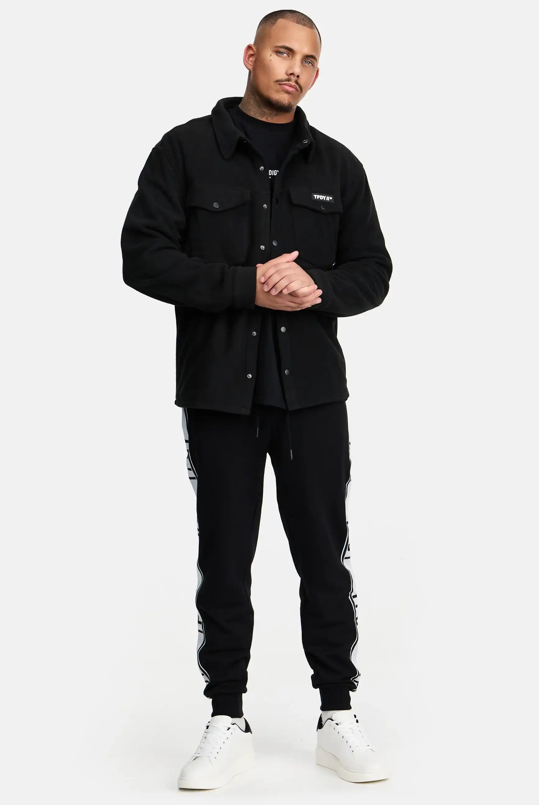 Ein Mann steht vor einem schlichten weißen Hintergrund und trägt das trueprodigy Bill Fleece Oversized Hemd in Schwarz über einem schwarzen Hemd, gepaart mit einer schwarzen Jogginghose mit weißen Seitenstreifen und weißen Sneakers. Er blickt nach vorne, die Hände leicht verschränkt.
