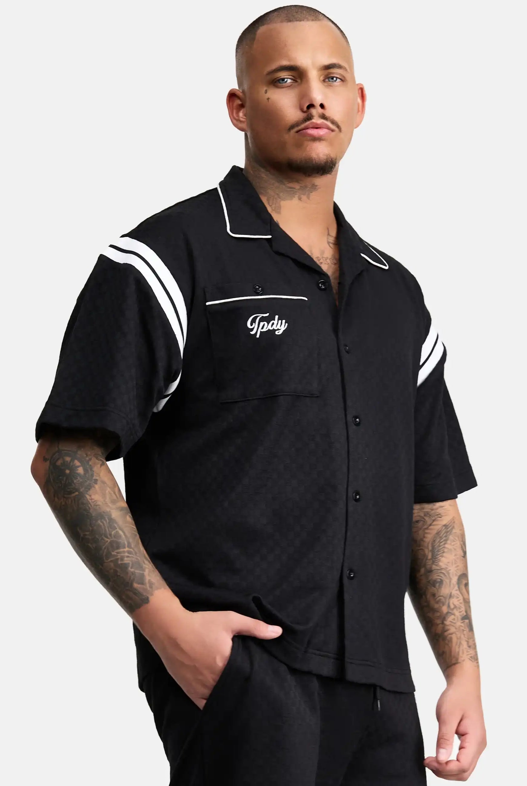 Ein Mann mit kurzen Haaren und Tattoos trägt das trueprodigy Steve Oversized Retro Hemd, ein schwarzes, kurzärmeliges Button-up-Hemd mit weißen Kontraststreifen und Tpdsy-Stickerei auf der Brusttasche, und steht mit einer Hand in der Tasche.
