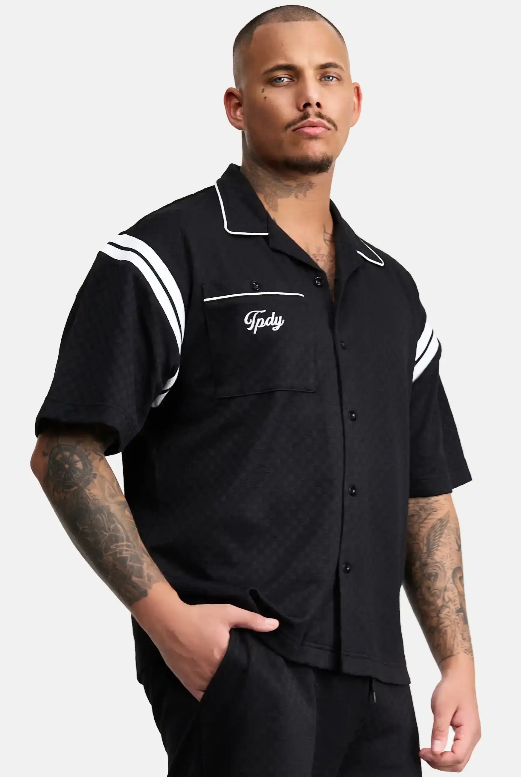 Ein Mann mit kurzen Haaren und Tattoos trägt das trueprodigy Steve Oversized Retro Hemd, ein schwarzes, kurzärmeliges Button-up-Hemd mit weißen Kontraststreifen und Tpdsy-Stickerei auf der Brusttasche, und steht mit einer Hand in der Tasche.