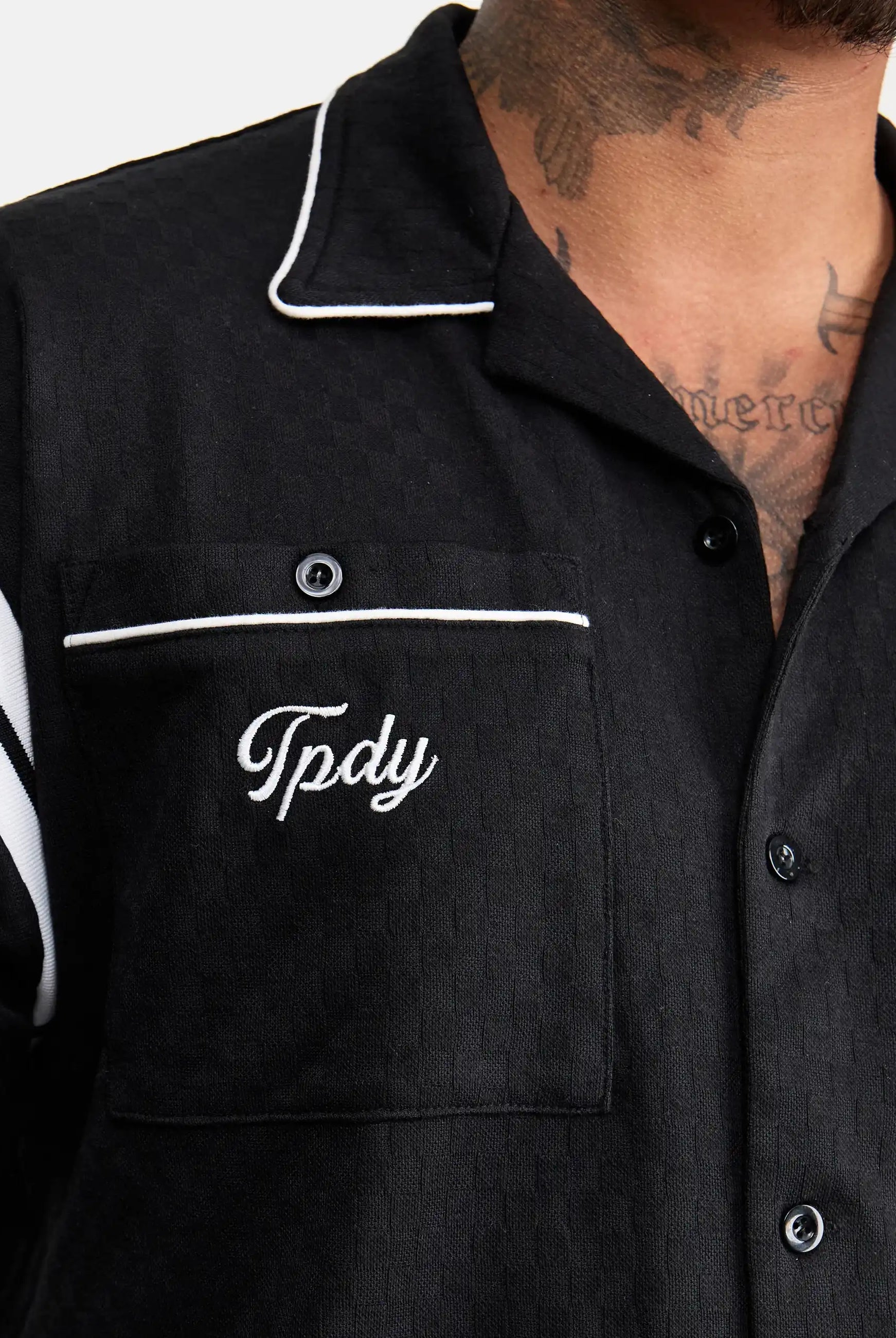 Eine Nahaufnahme zeigt eine Person, die das trueprodigy Steve Oversized Retro Hemd mit weißen Kontraststreifen an Kragen und Tasche trägt. Auf der Brusttasche ist "Tpdy" aufgestickt, und auf der oberen Brust und im Nacken ist teilweise ein Tattoo zu sehen.