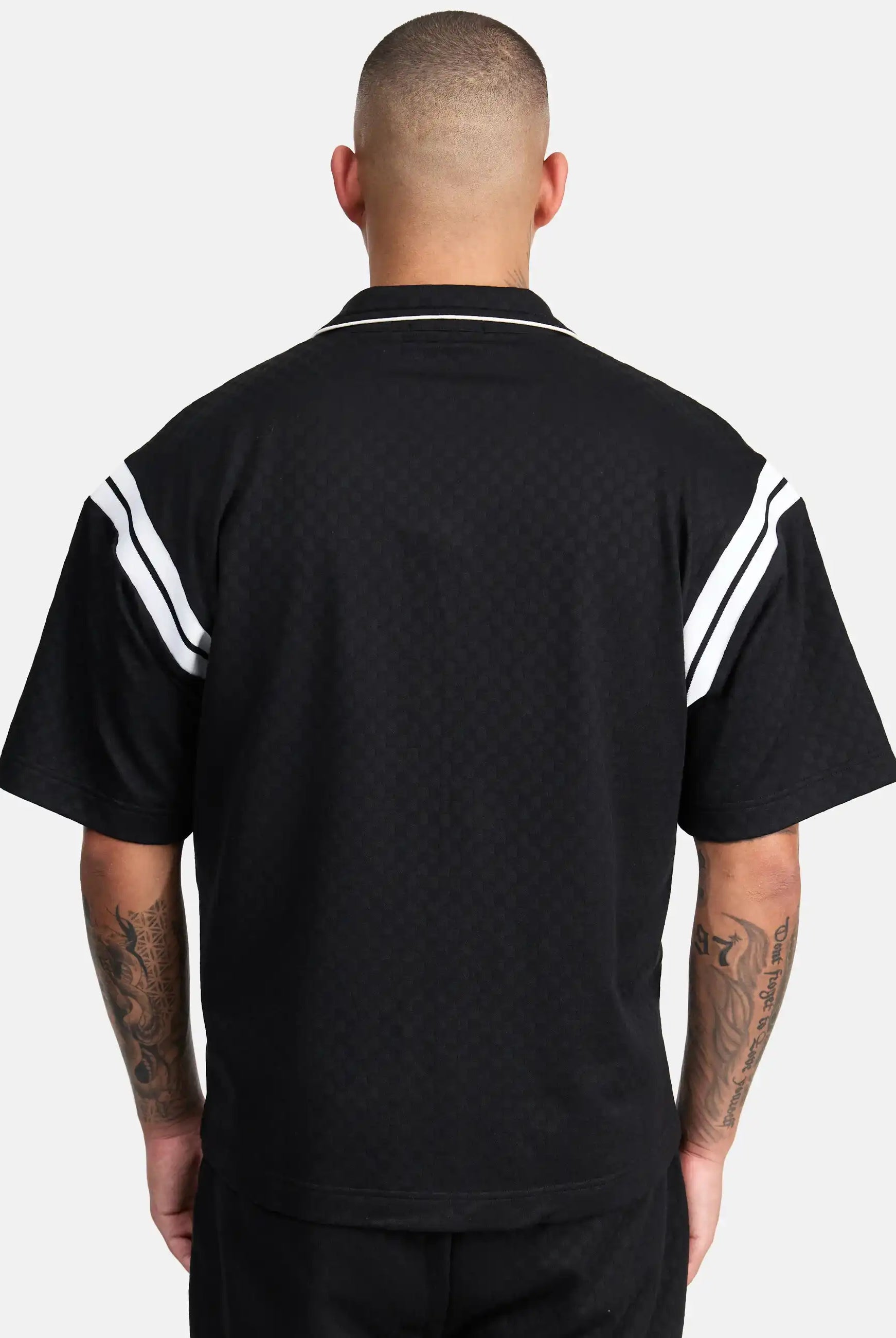 Von hinten ist eine Person mit kurzen Haaren zu sehen, die das trueprodigy Steve Oversized Retro Hemd in Schwarz mit weißen Kontraststreifen an den Schultern trägt. Ihre tätowierten Arme sind sichtbar, während sie vor einem schlichten hellen Hintergrund stehen.