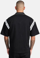 Von hinten ist eine Person mit kurzen Haaren zu sehen, die das trueprodigy Steve Oversized Retro Hemd in Schwarz mit weißen Kontraststreifen an den Schultern trägt. Ihre tätowierten Arme sind sichtbar, während sie vor einem schlichten hellen Hintergrund stehen.