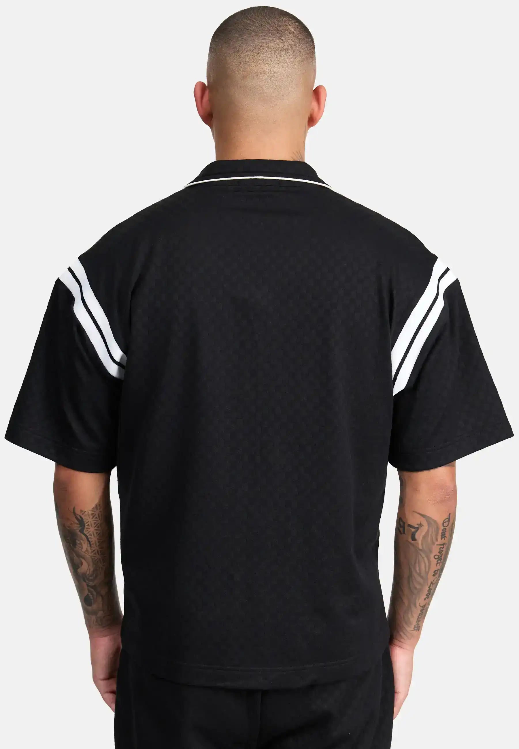 Von hinten ist eine Person mit kurzen Haaren zu sehen, die das trueprodigy Steve Oversized Retro Hemd in Schwarz mit weißen Kontraststreifen an den Schultern trägt. Ihre tätowierten Arme sind sichtbar, während sie vor einem schlichten hellen Hintergrund stehen.