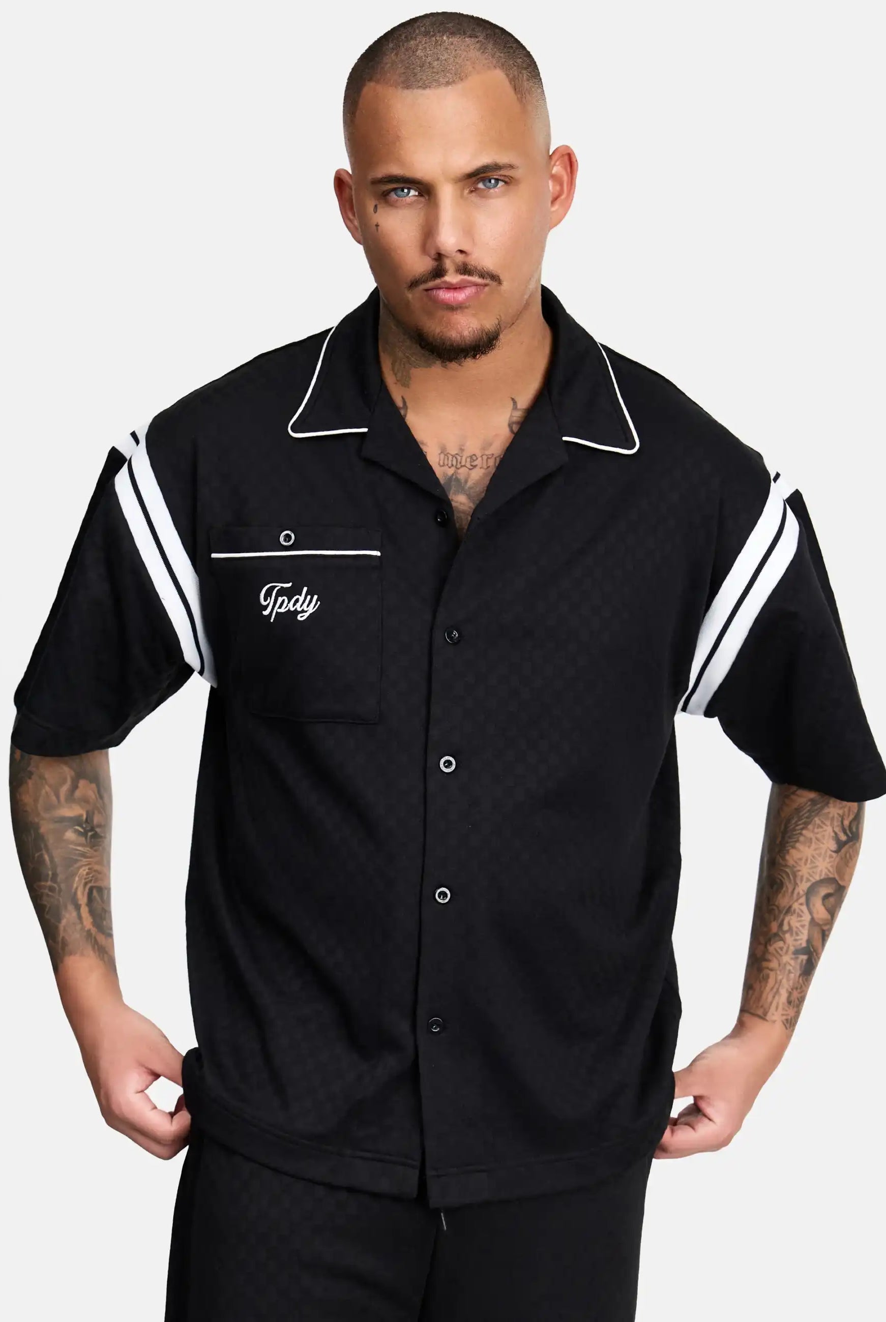 Ein tätowierter Mann mit Kurzhaarschnitt trägt das trueprodigy Steve Oversized Retro Hemd, ein schwarzes, kurzärmeliges Button-up-Hemd mit weißen Kontraststreifen an den Ärmeln, weißer Paspelierung am Kragen und gesticktem "Tpdv" auf der Brusttasche.