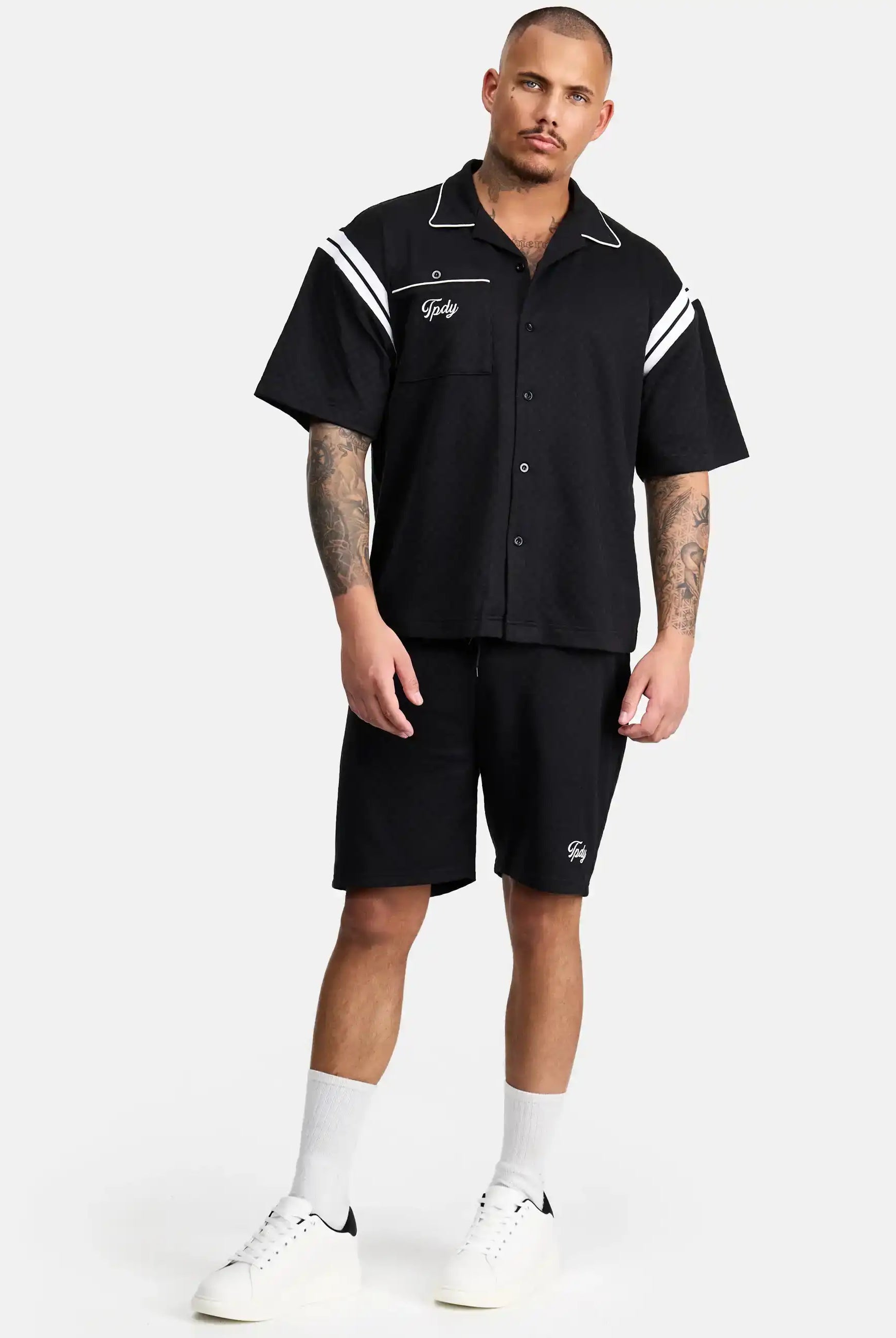 Ein Mann mit Tattoos und Kurzhaarschnitt steht vor einem schlichten Hintergrund und trägt das Steve Oversized Retro Hemd von trueprodigy mit weißen Kontraststreifen an den Schultern, schwarze Shorts, weiße Socken und Turnschuhe.